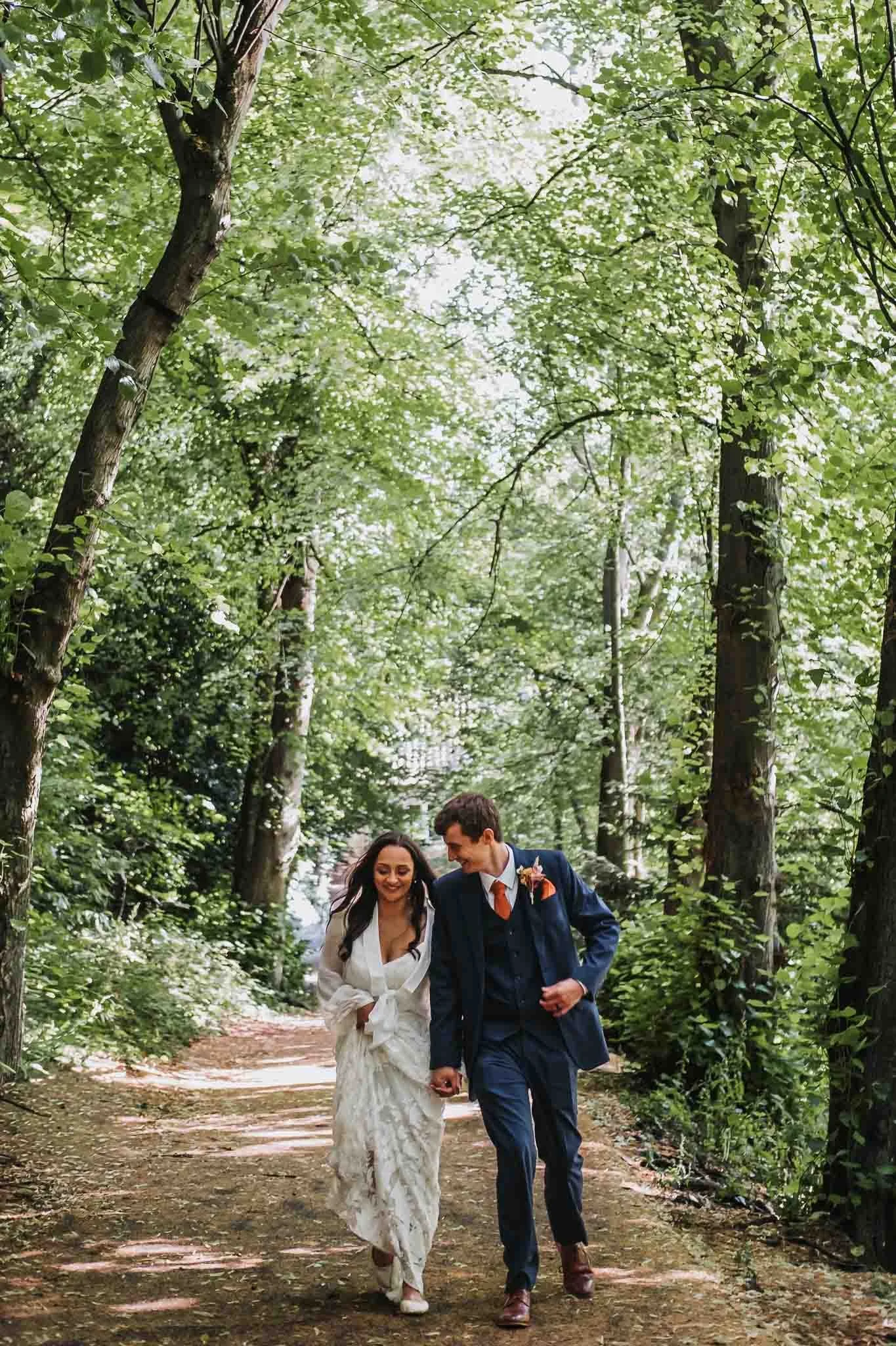 A couple dressed in wedding attire walking hand-in-hand along a wooded forest trail, smiling and looking at each other, with tall trees and lush green foliage surrounding them in Durham.