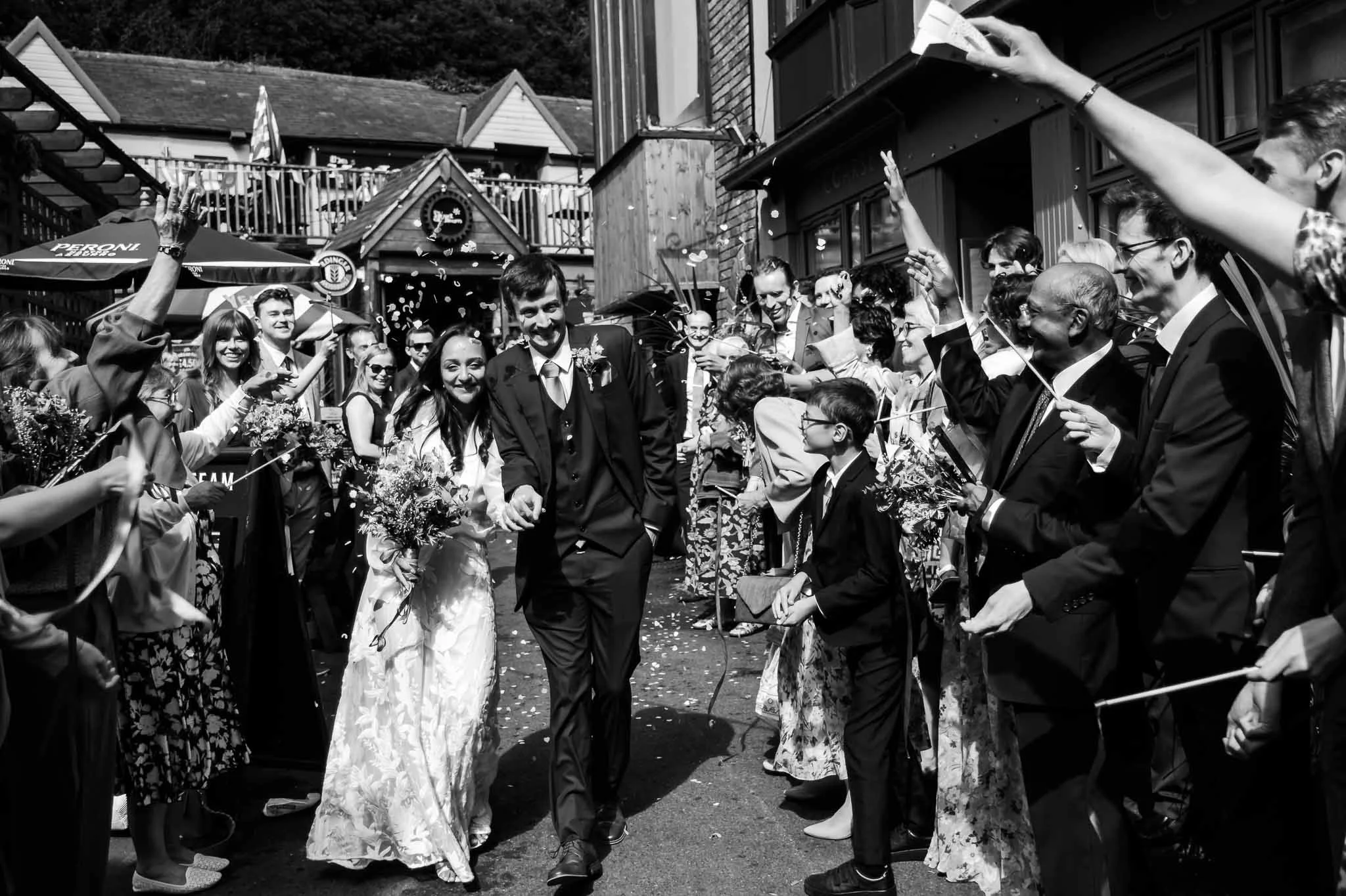 Black and white photo of a wedding celebration with a bride and groom walking through a crowd holding confetti and flowers, surrounded by smiling guests.