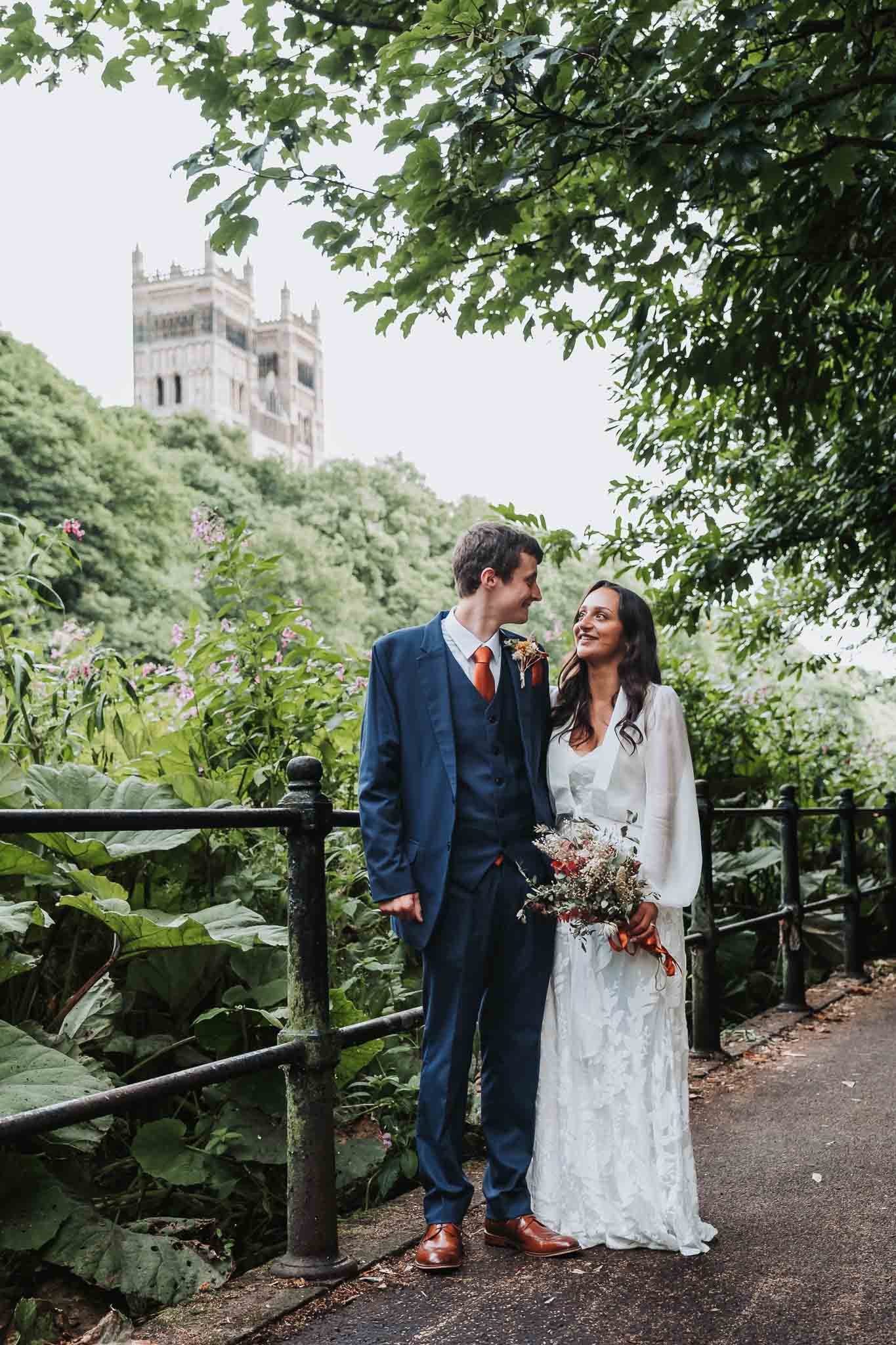 A couple dressed in wedding attire, standing on a park pathway surrounded by lush greenery, with a historic building visible in the background.