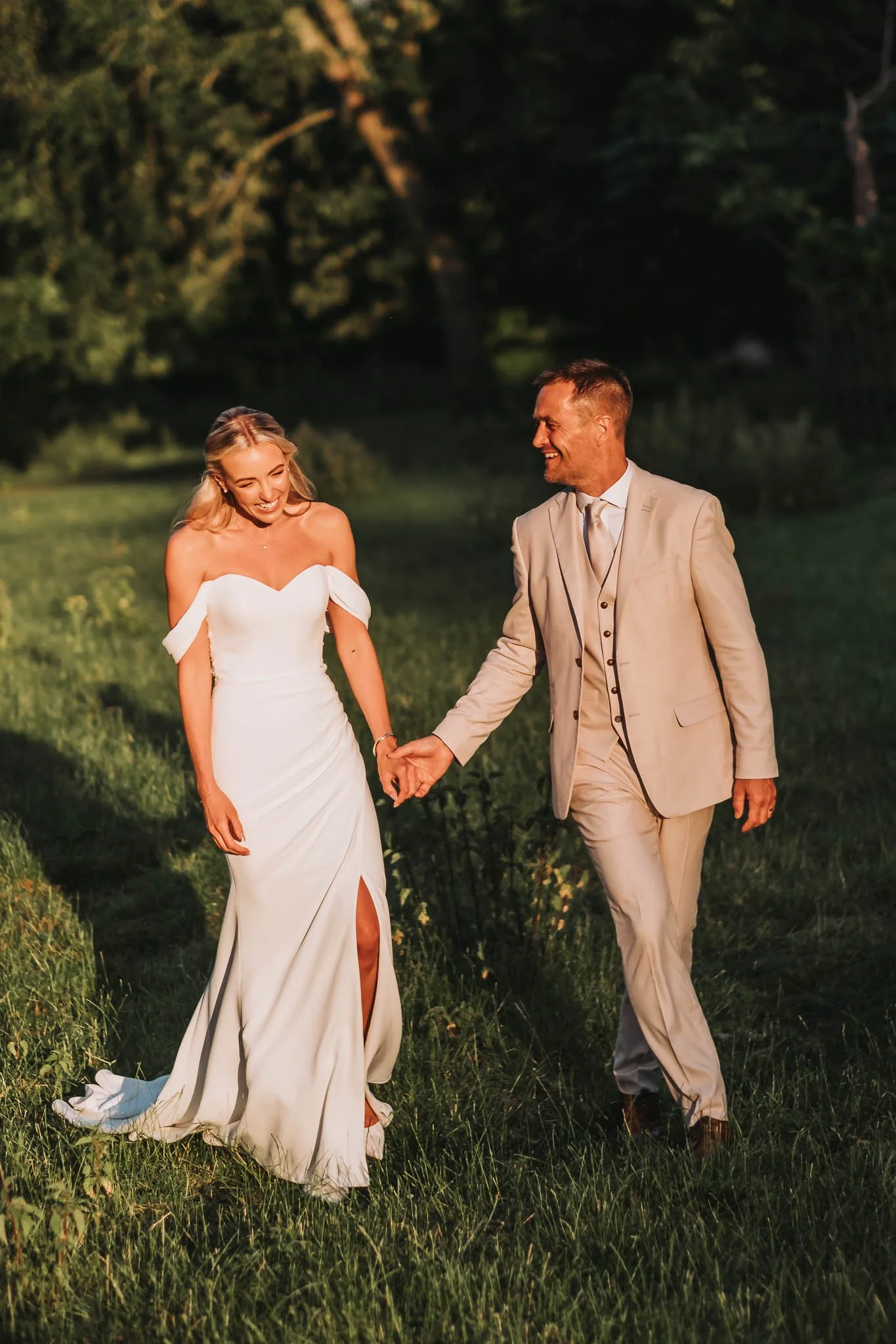 A bride and groom holding hands and walking outdoors on a grassy path during sunset, with trees in the background in Whitworth Hall.