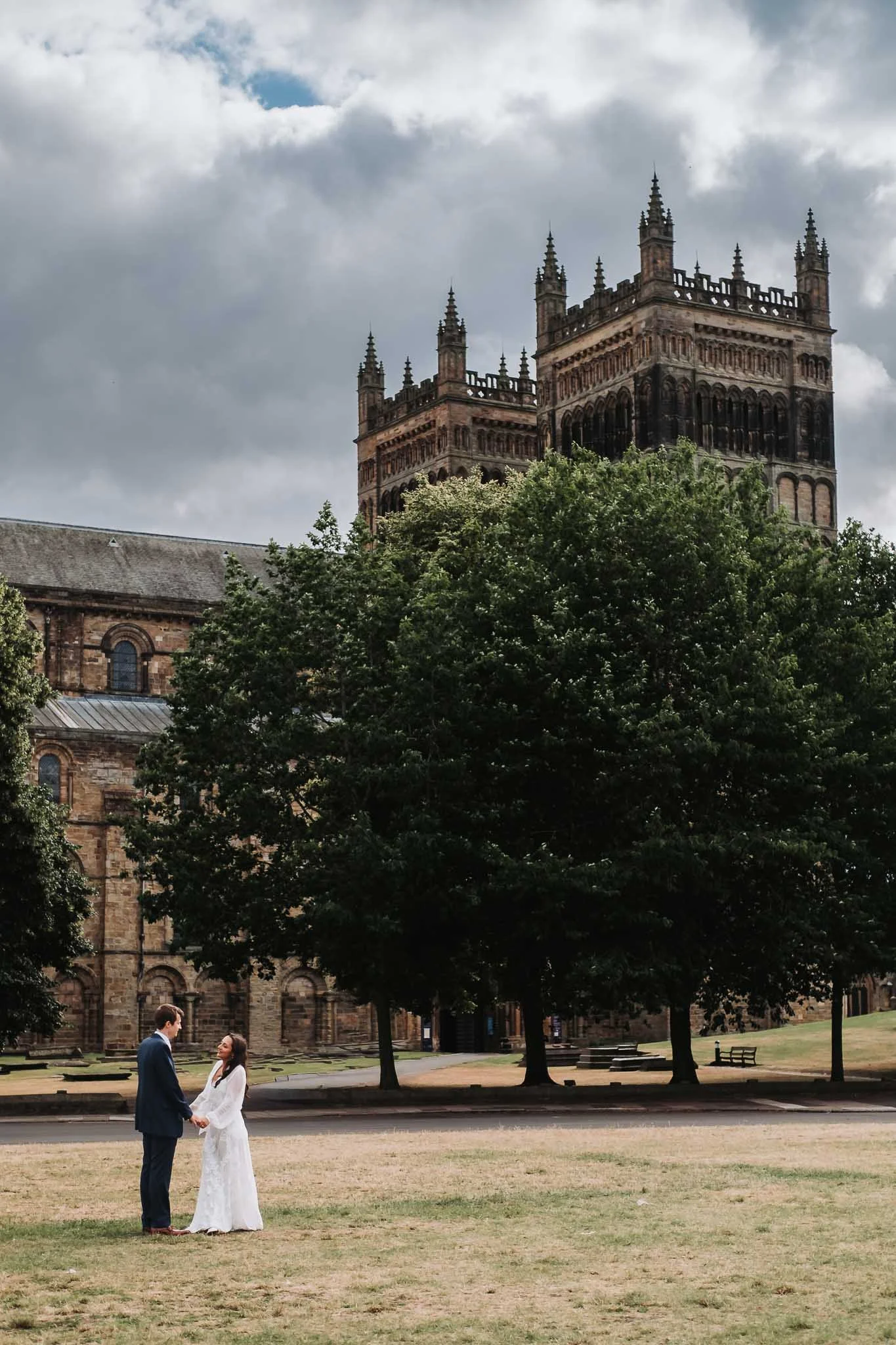 A bride and groom holding hands, standing outdoors in front of an old stone church with Gothic architecture, surrounded by trees under a cloudy sky.