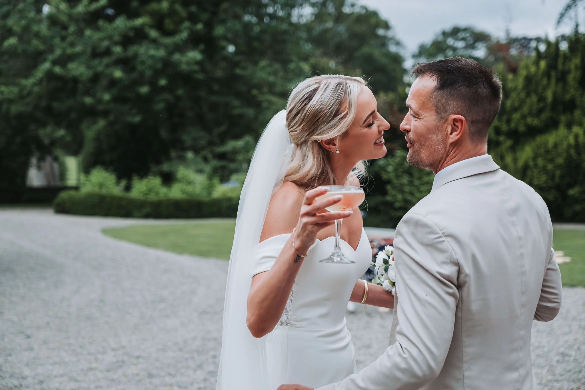 A bride and groom smiling at each other outdoors, the bride holding a glass of pink drink, surrounded by lush greenery.