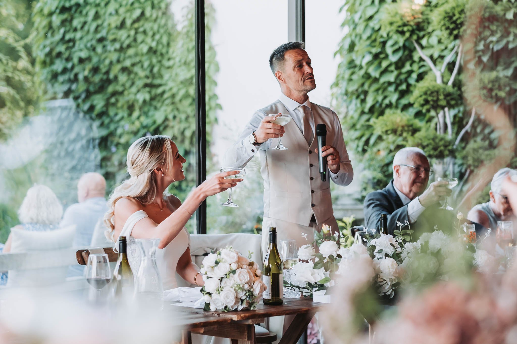 A man in a white suit giving a speech at a wedding reception, holding a glass of wine and a microphone, surrounded by guests at a decorated table with flowers.