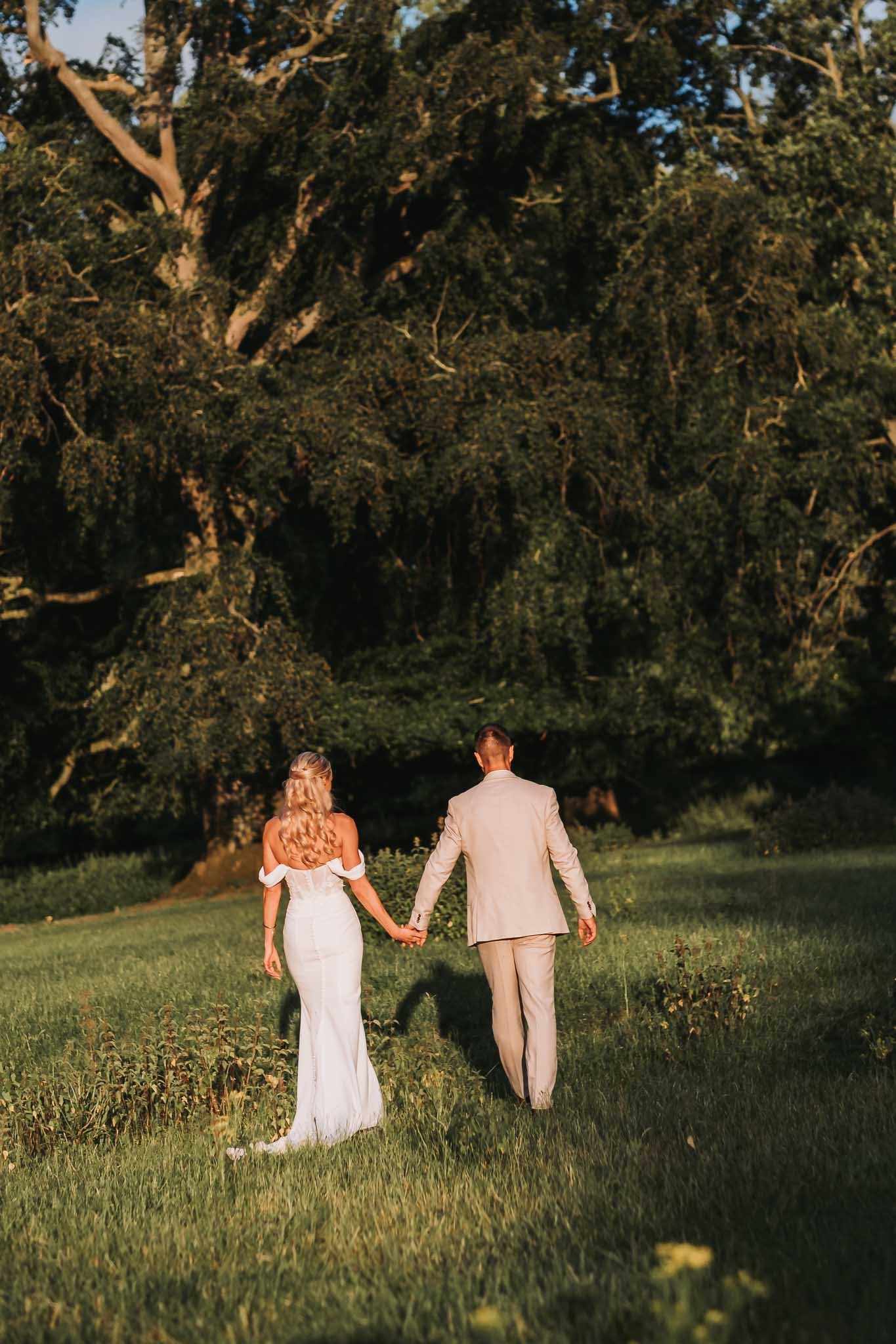 A couple dressed in wedding attire walking hand in hand through a grassy field towards a large tree at Whitworth Hall, with the sun setting behind them.