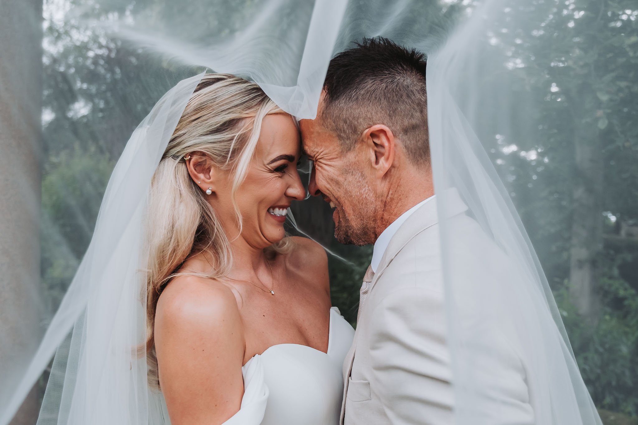A bride and groom are face to face, touching foreheads with eyes closed, smiling. The bride has blonde hair and is wearing a strapless white wedding dress and a veil. They are outdoors with trees in the background at Whitworth Hall.