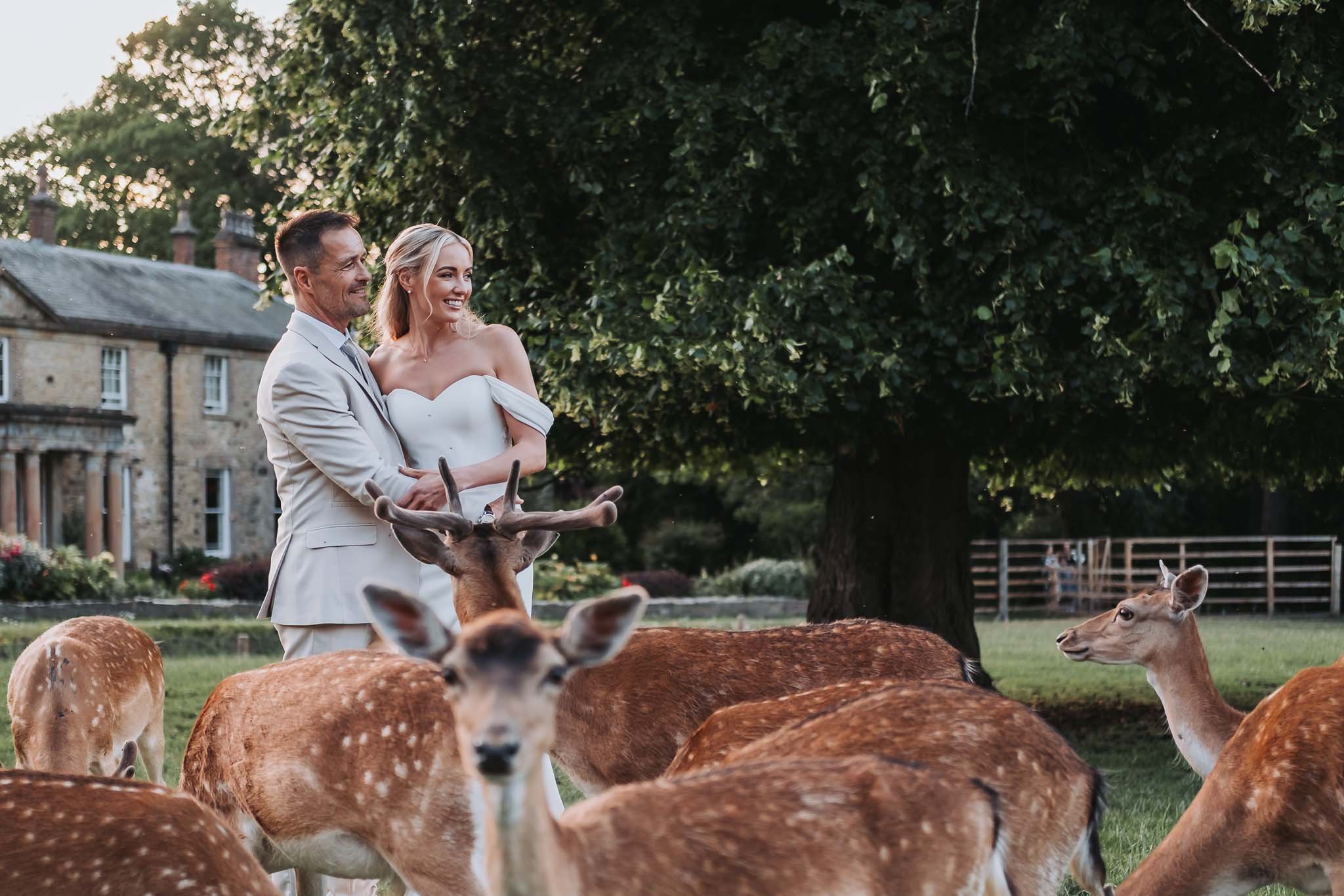A man and woman in formal attire standing among deer outdoors with green trees and Whitworth Hall in the background.