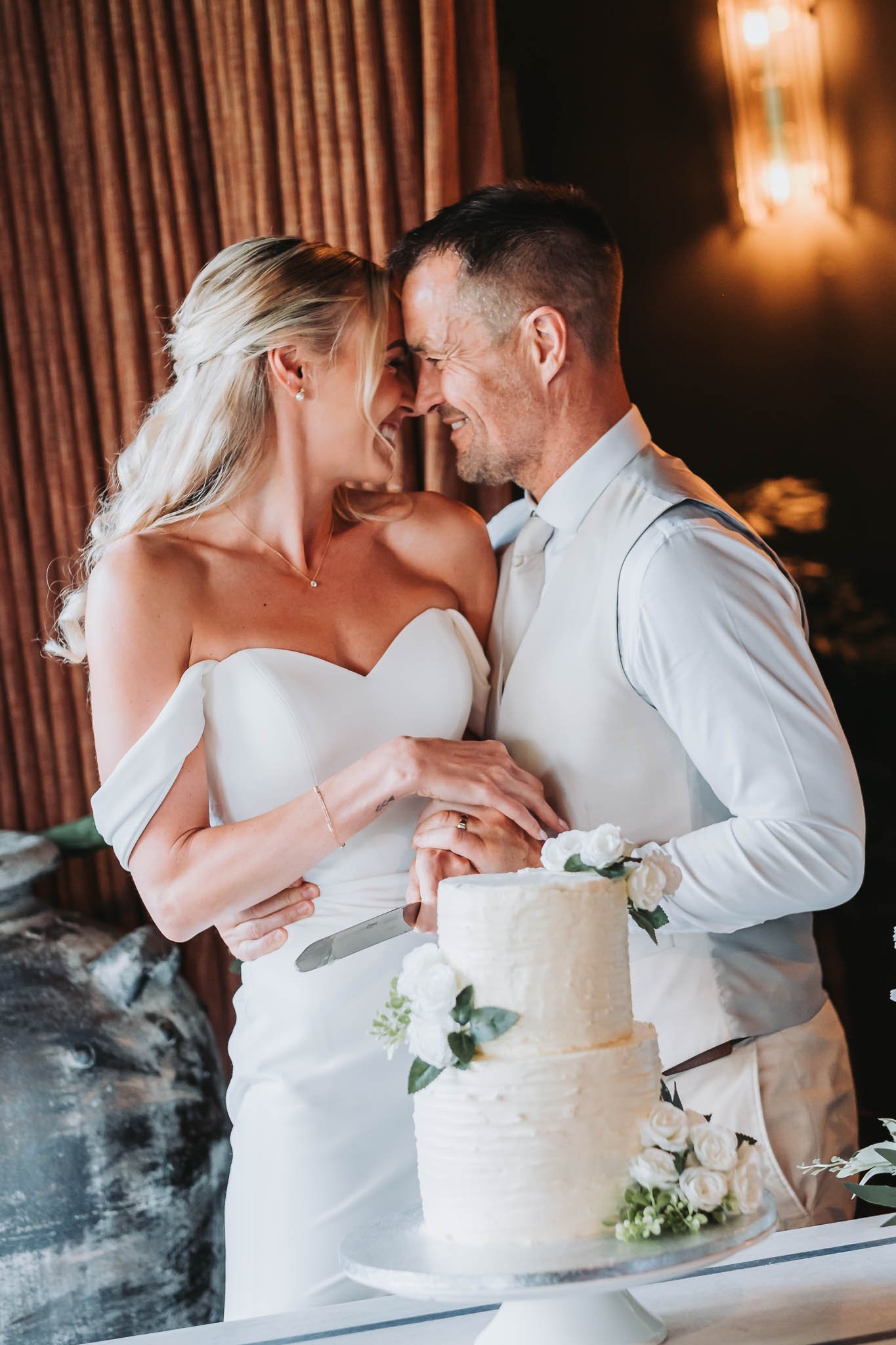 A bride and groom are smiling and touching foreheads with their eyes closed during a wedding cake cutting ceremony at Whitworth Hall. The bride is wearing a white off-shoulder dress and the groom is in a white shirt.