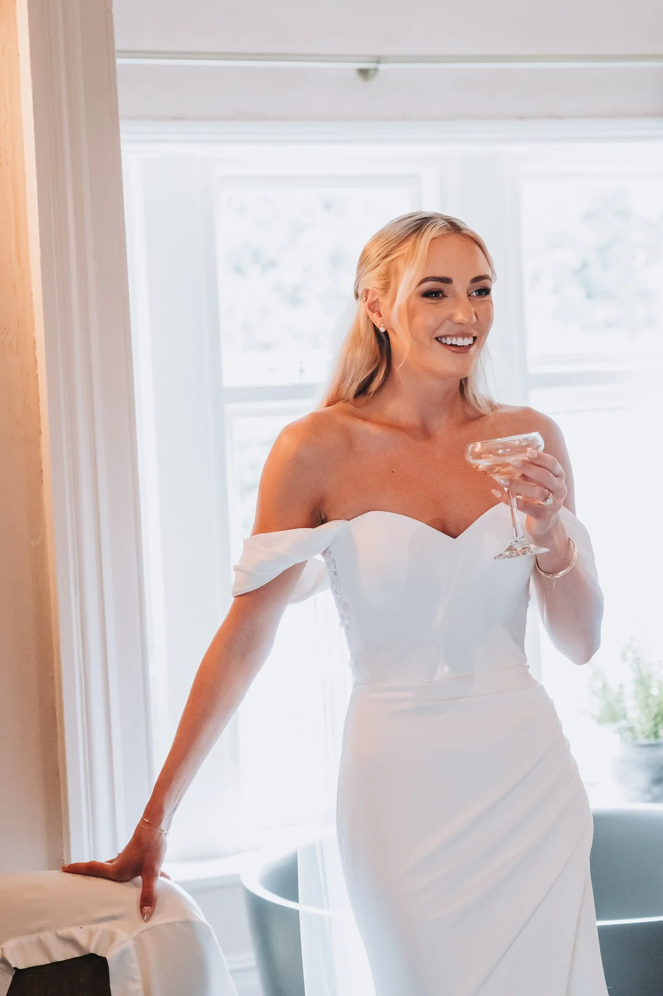 A woman in a white dress holding a glass of champagne, smiling, standing by a window with sunlight coming through, in a bright room.