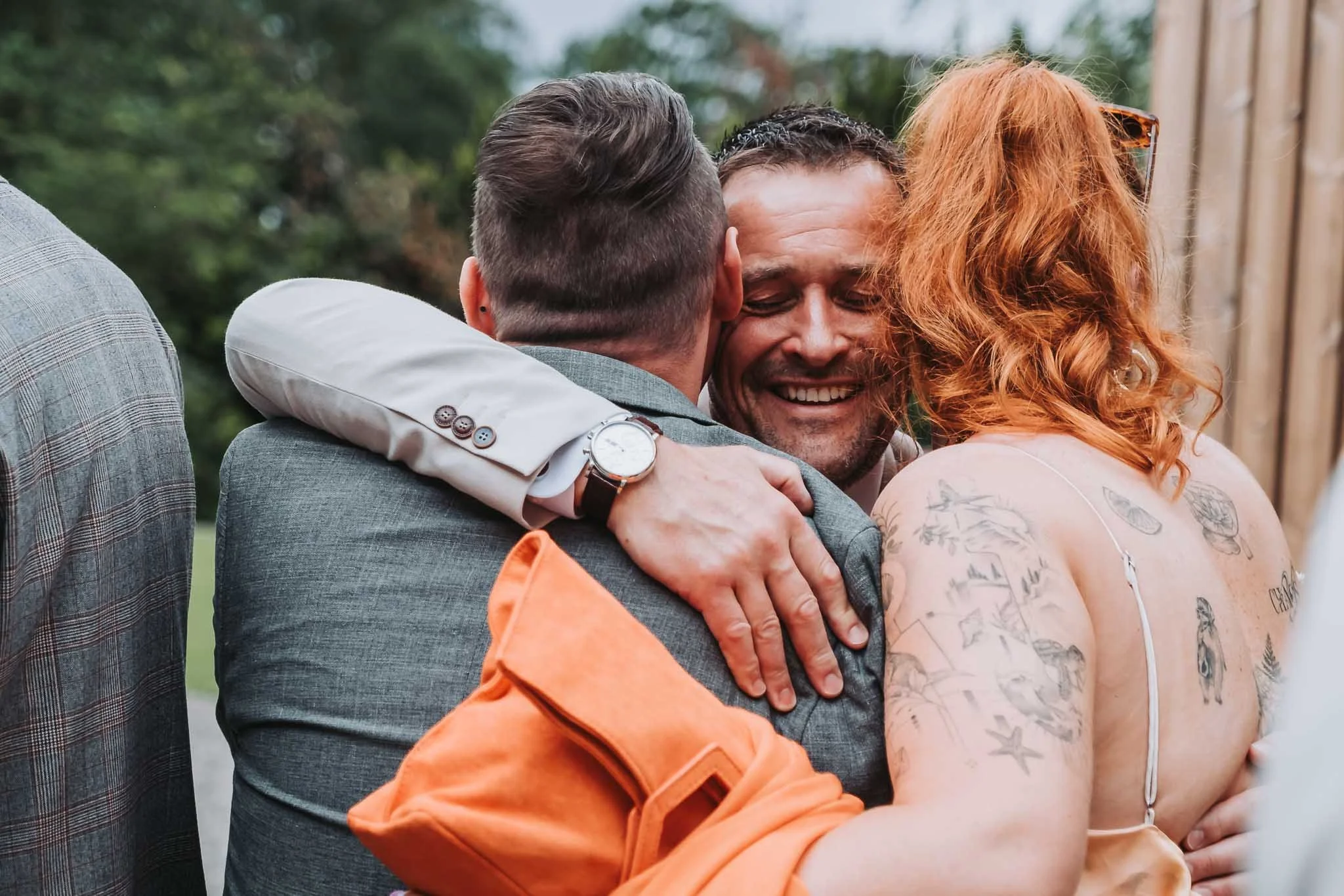 Three people sharing a group hug outdoors. One person has tattoos on their arm and red hair, another has dark hair and a gray suit, and the third has short hair and wears glasses. They appear happy and close.