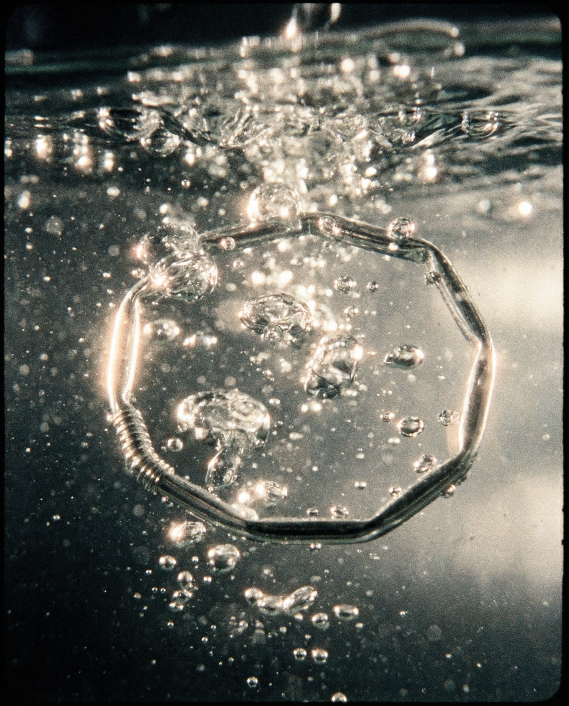 Ein silberner Armreif, der wie ein geknickter Plastiktrinkhalm aussieht, in Wasser, umgeben von Blasen, die vom Wasser aufsteigen.