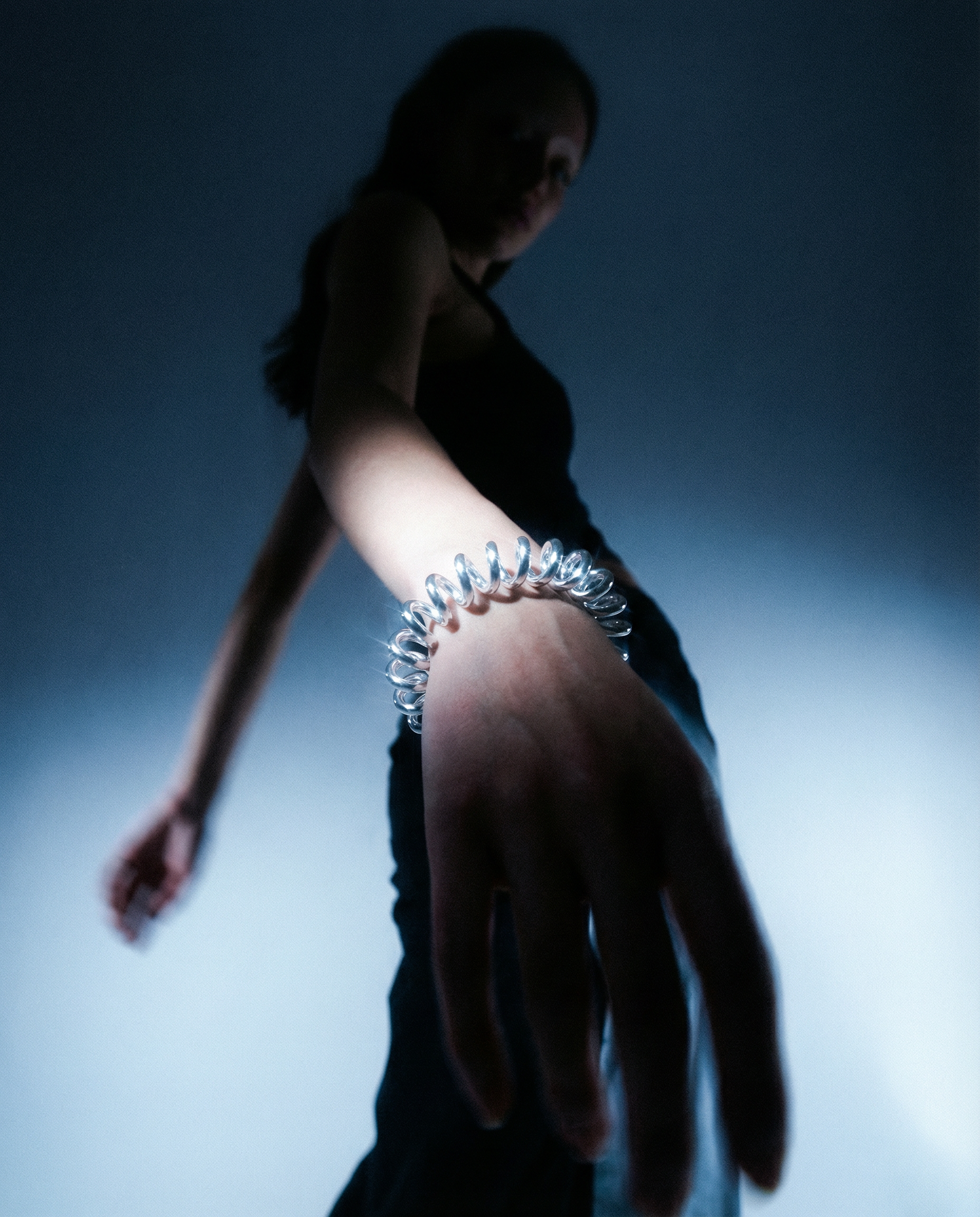 Junge Frau in dunklem Top zeigt auf die Kamera, trägt einen silbernen, spiralförmigen Schmuck am Handgelenk, im Hintergrund unscharf, dunkler Ton.