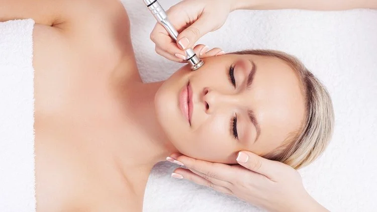 Woman receiving a facial treatment with a small tool near her face, lying down with eyes closed.