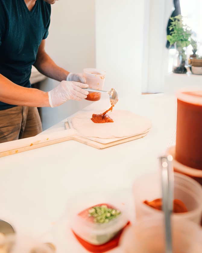 A person wearing gloves spreading sauce onto a piece of dough on a marble surface, with various containers of ingredients nearby and a plant on a windowsill in the background.