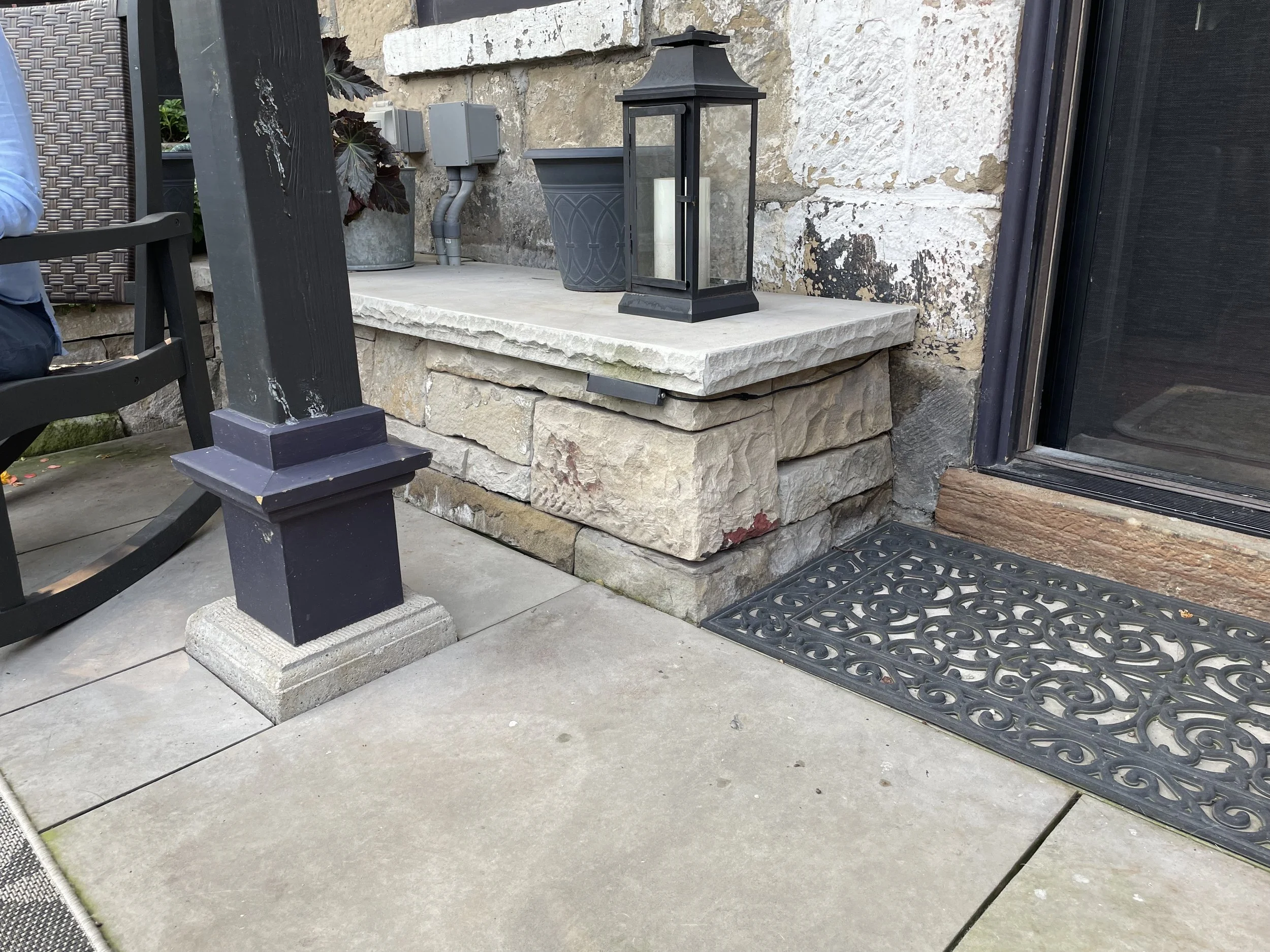 Close-up of a porch step built with large beige stone blocks, with a black lantern and potted plants on top. A black decorative doormat is in front of a sliding glass door.