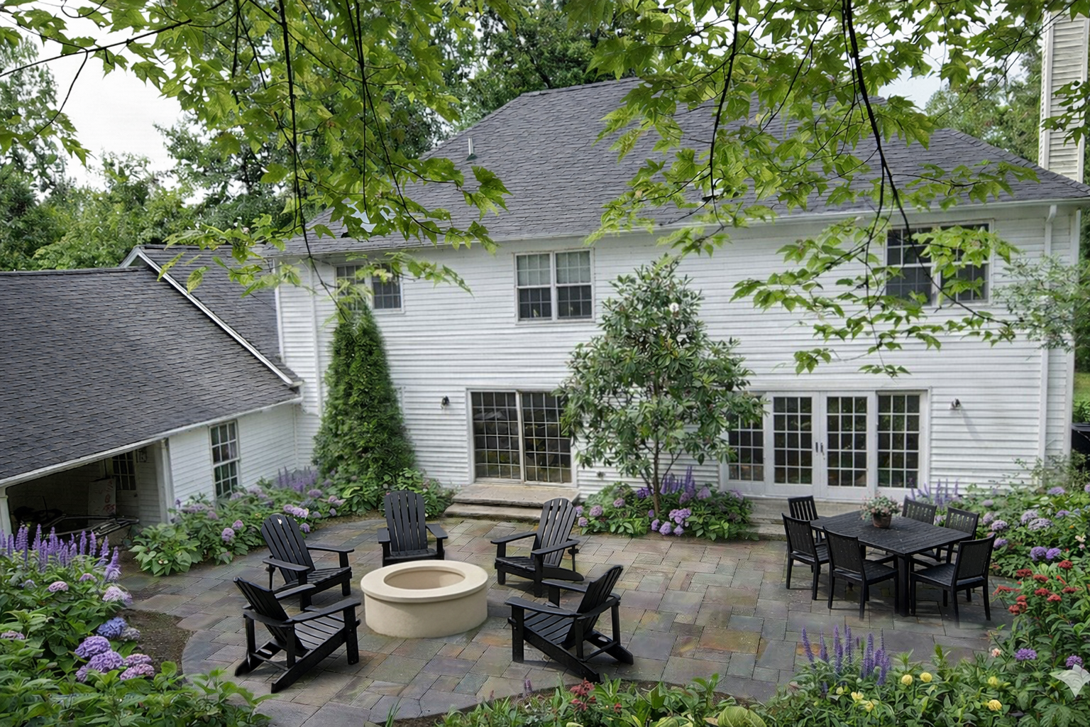 Backyard patio with a fire pit surrounded by four Adirondack chairs, adjacent to a garden with purple and green plants, and a white house with large glass doors and multiple windows.