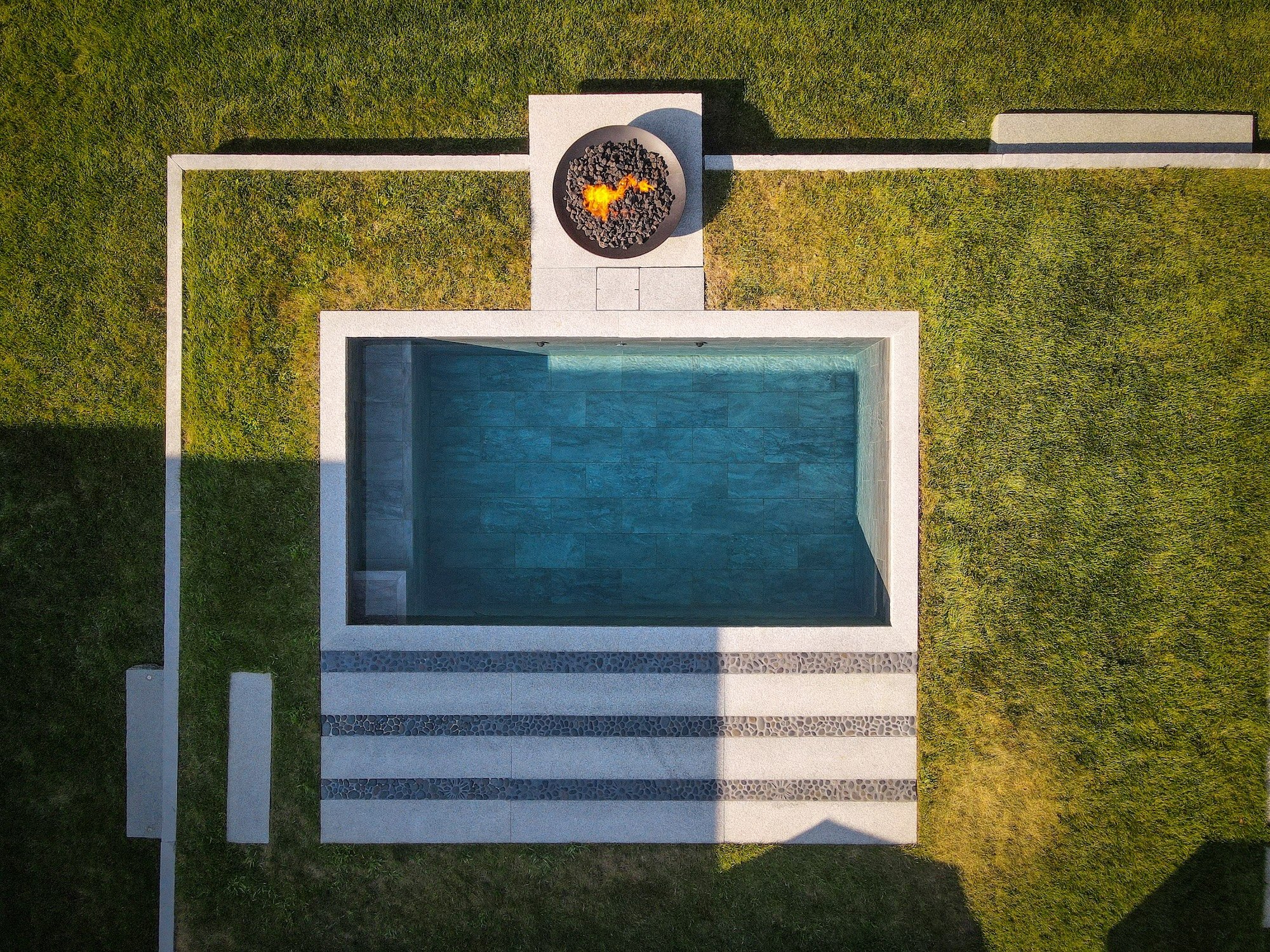 A modern swimming pool with blue water, surrounded by white concrete and decorative stone borders, located on a grassy lawn with a fire pit nearby.