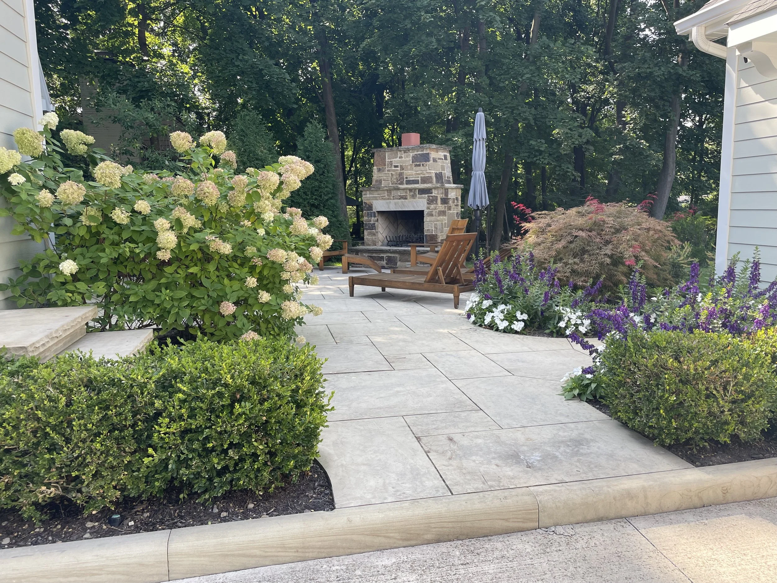 A backyard patio with a stone fireplace, wooden chairs, and a large outdoor umbrella surrounded by various flowering plants and shrubs, with a wooded area in the background.