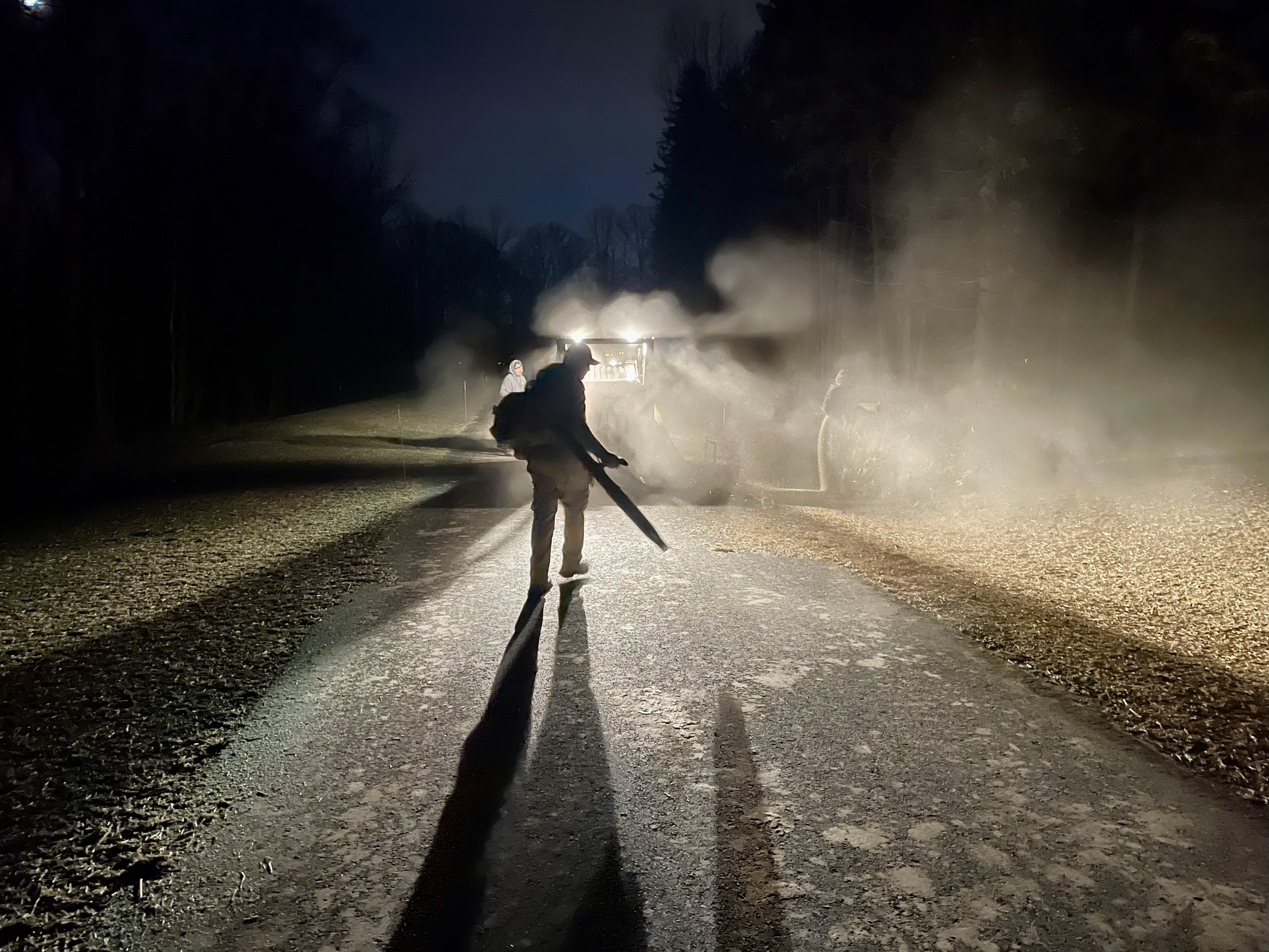 Firefighter using a hose to spray water on a fire on a rural road at night, with smoke and mist in the air and trees on both sides.