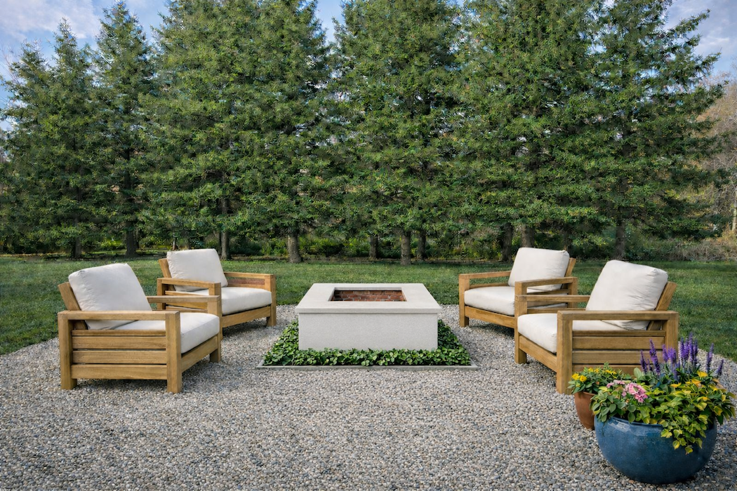 Outdoor patio with four wooden armchairs with white cushions arranged around a fire pit, with a large green tree line in the background and potted flowers to the side.