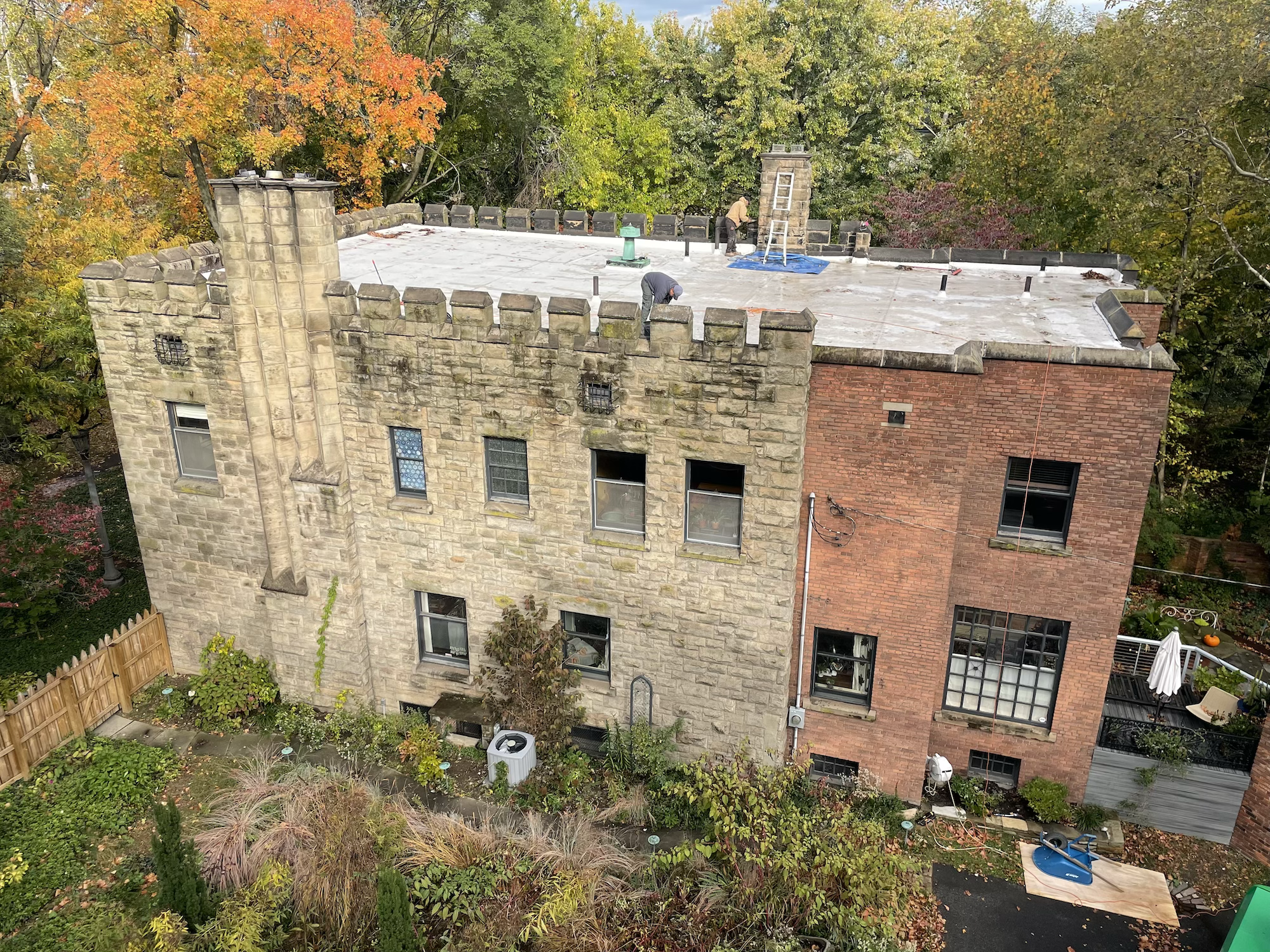 Two workers standing on a flat rooftop with a ladder, working on the roof of a brick building surrounded by trees with autumn foliage.