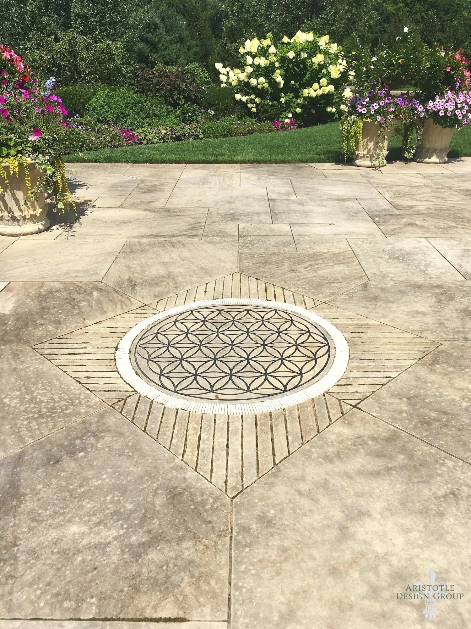 Decorative circular drain cover on a concrete patio surrounded by patterned tiles, with flower pots and a garden with colorful flowers in the background.