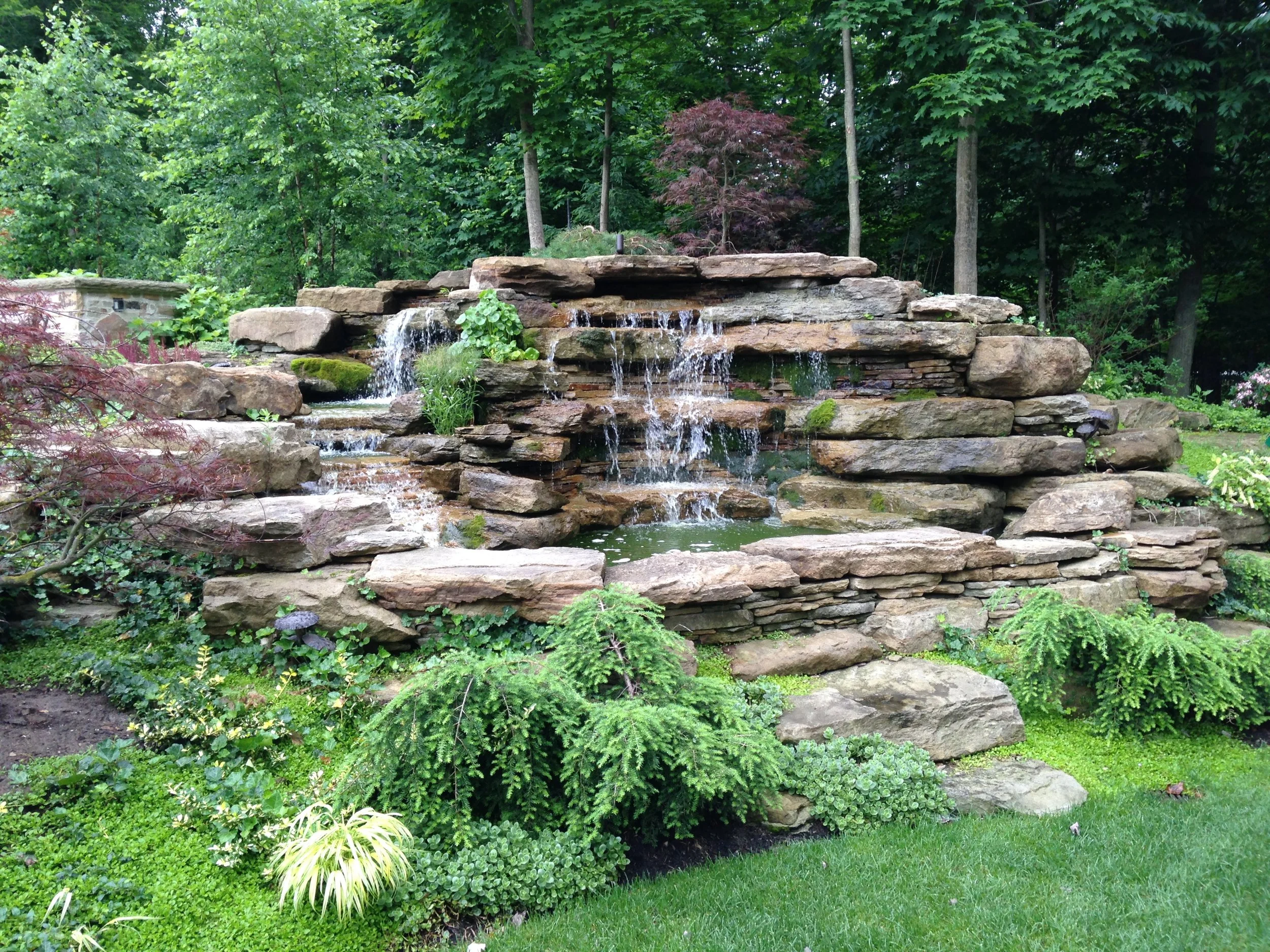 A landscaped garden with a tiered stone waterfall feature surrounded by various green plants and trees.