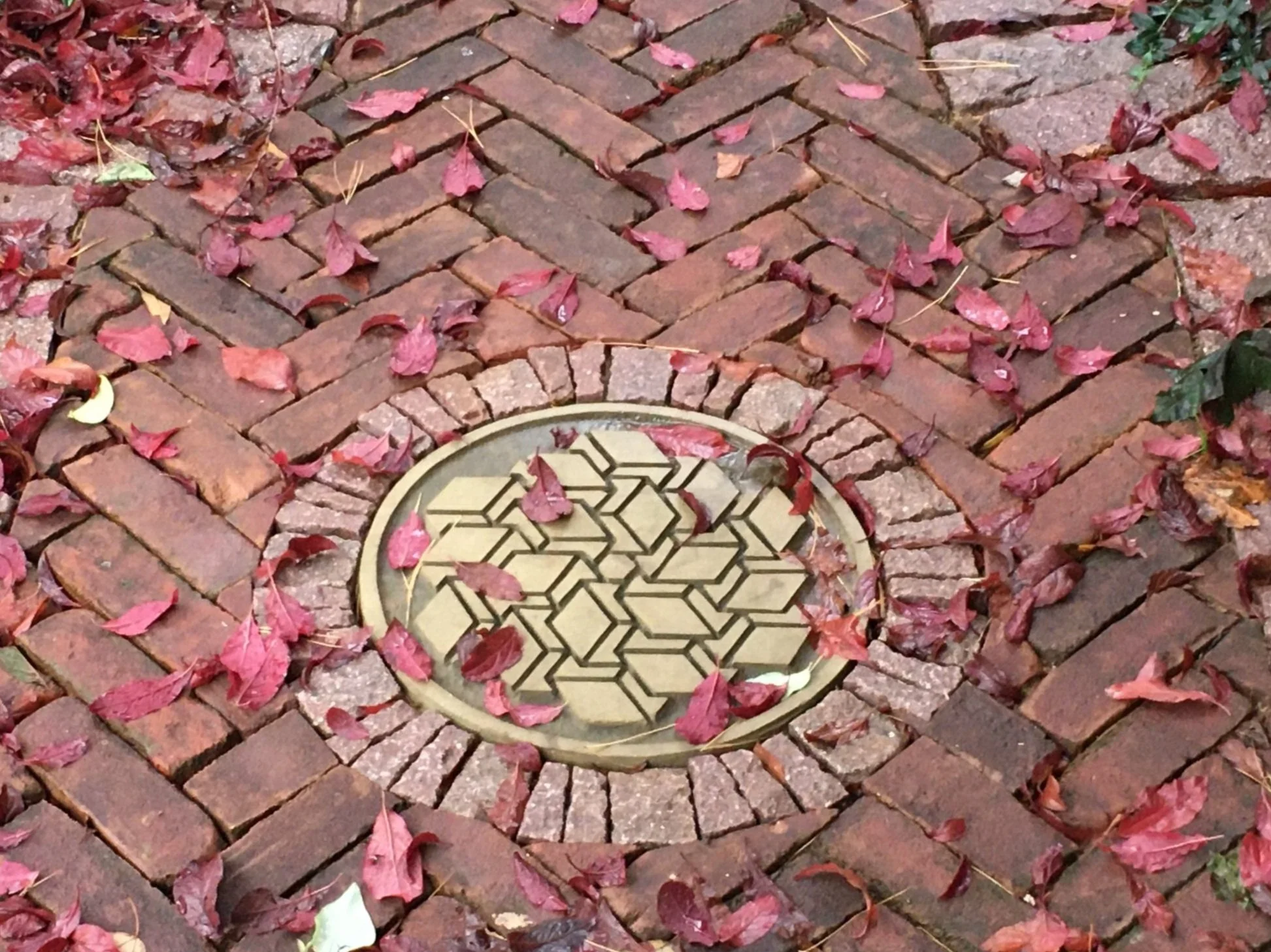 A brick sidewalk with fallen red and pink leaves surrounding a round decorative metal cover with geometric patterns.