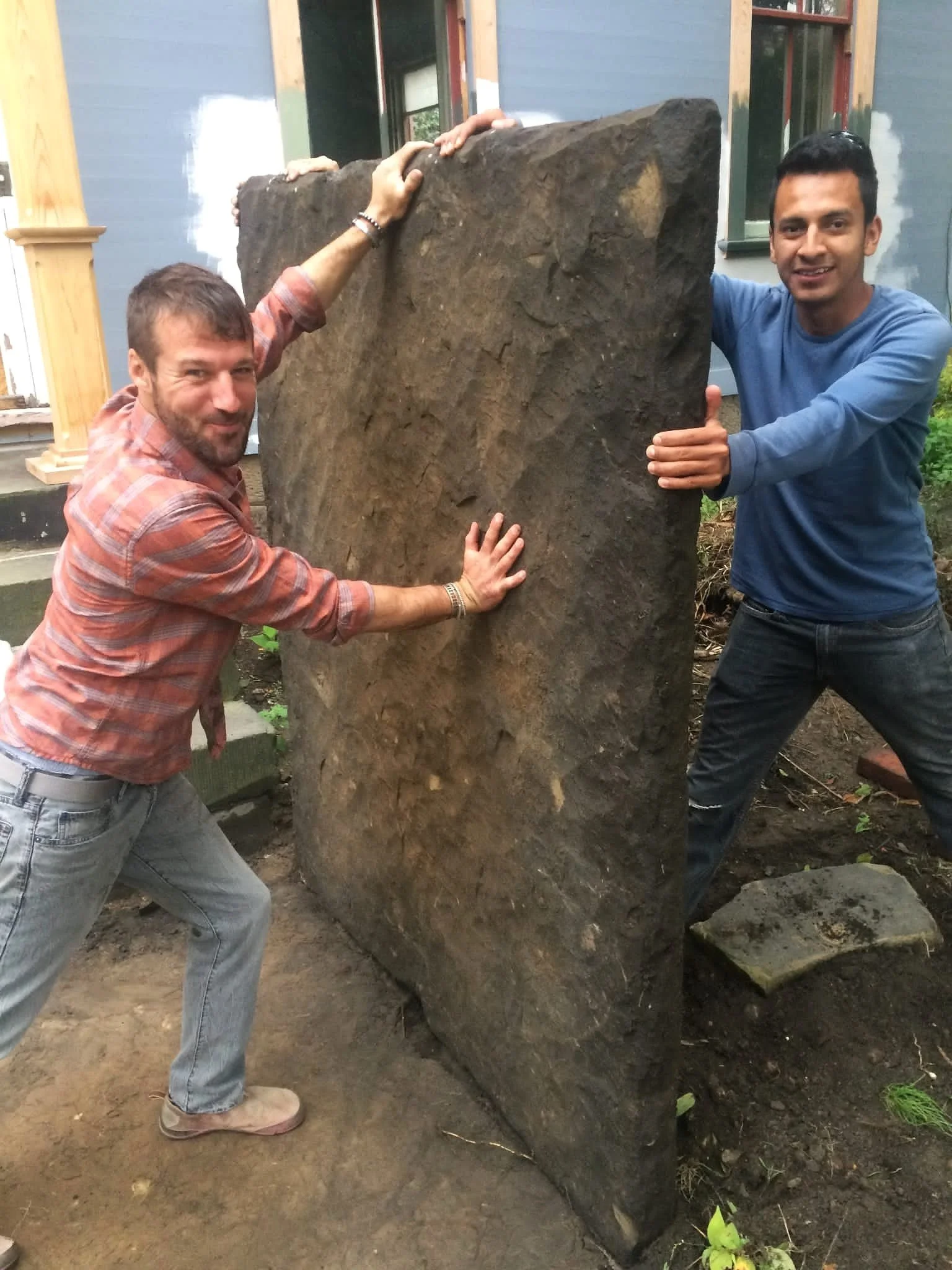 Two men lifting and holding a large rectangular stone slab outdoors near a house, with one man in a plaid shirt and the other in a blue shirt.