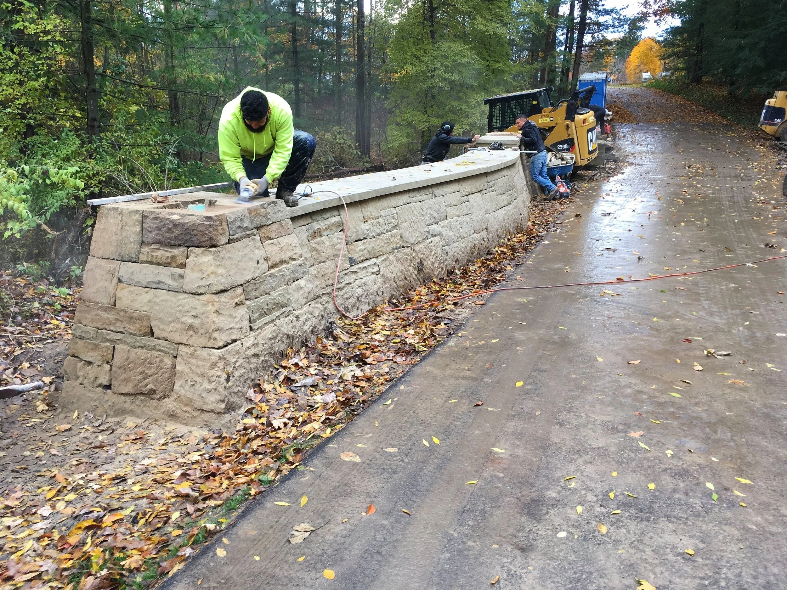 Workers building a stone retaining wall along a wet, leaf-covered road in a wooded area during autumn.