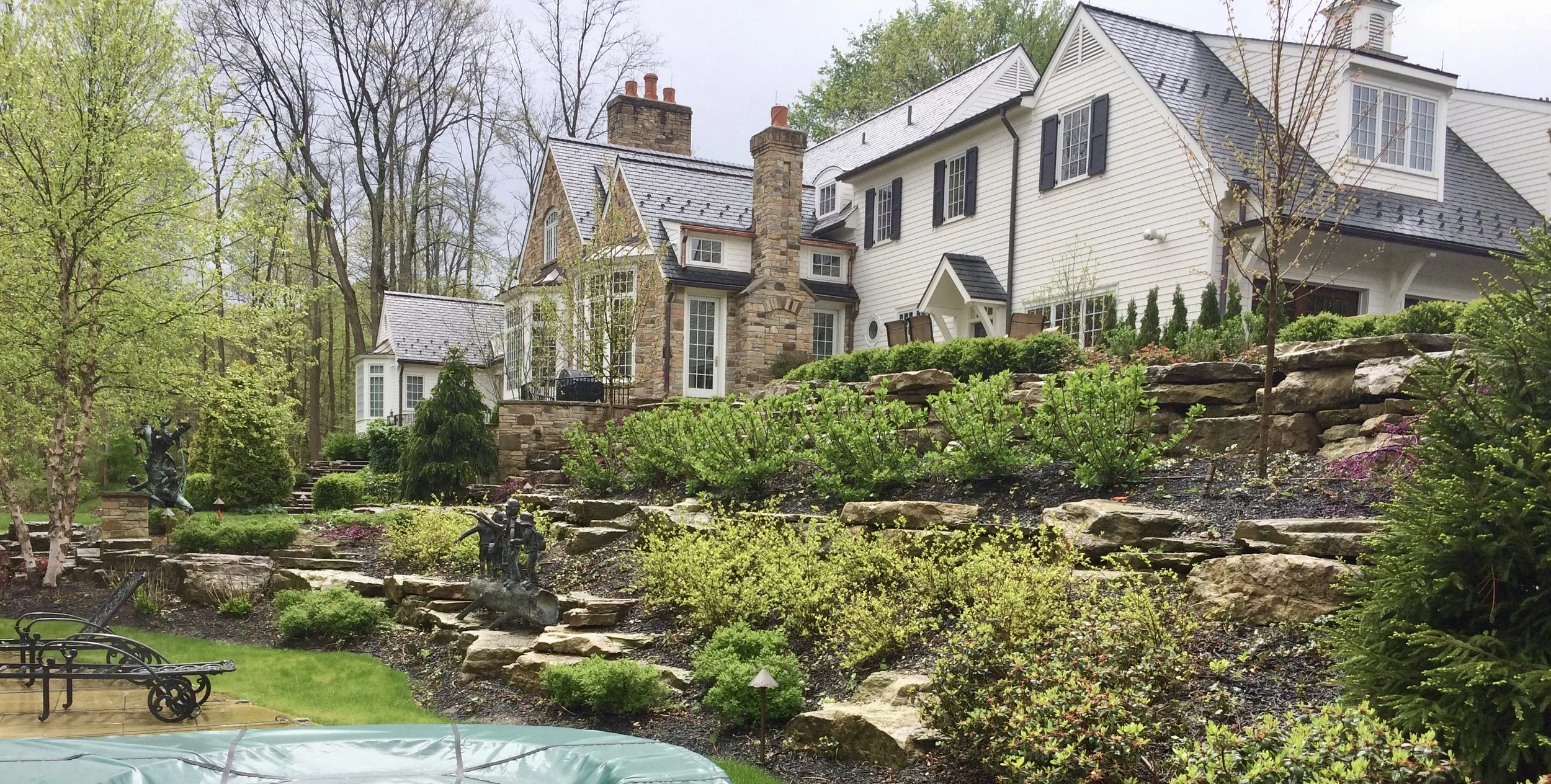 A large multi-story house with a stone and white siding exterior, surrounded by a landscaped garden with various trees, shrubs, stone steps, and decorative statues.