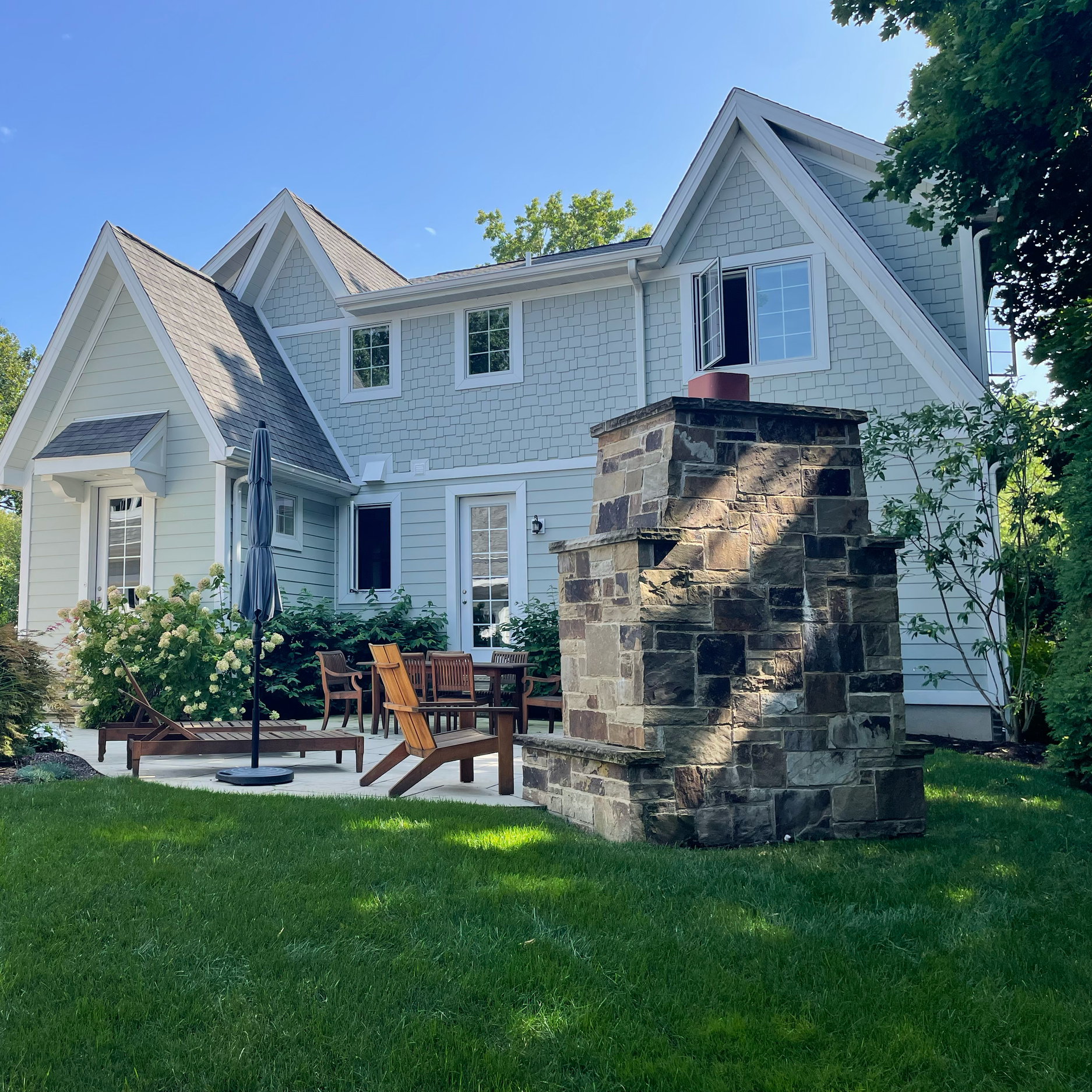 Backyard patio with outdoor furniture, deck chairs, an umbrella, blooming bushes, a stone firepit, and a white two-story house with multiple windows and a chimney, on a sunny day with a clear sky.