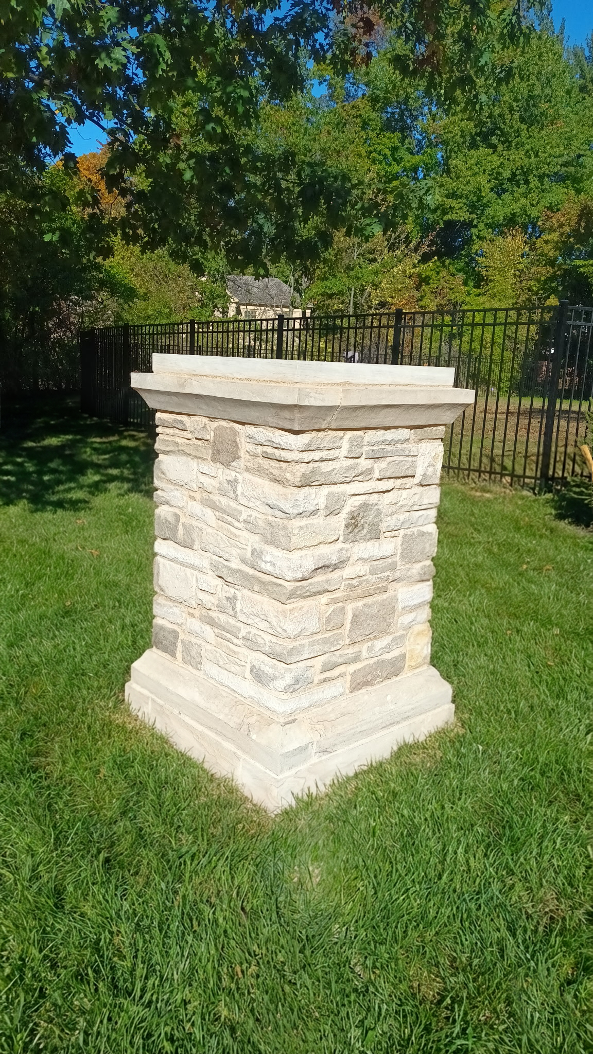 A stone and brick pedestal with layered capstone in a grassy yard, surrounded by trees and a black metal fence in the background.