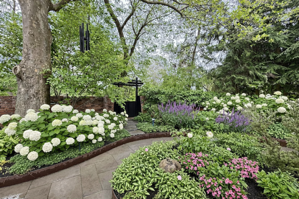 A lush garden with a winding concrete pathway, surrounded by blooming white, pink, and purple flowers, large trees, and dense green foliage.