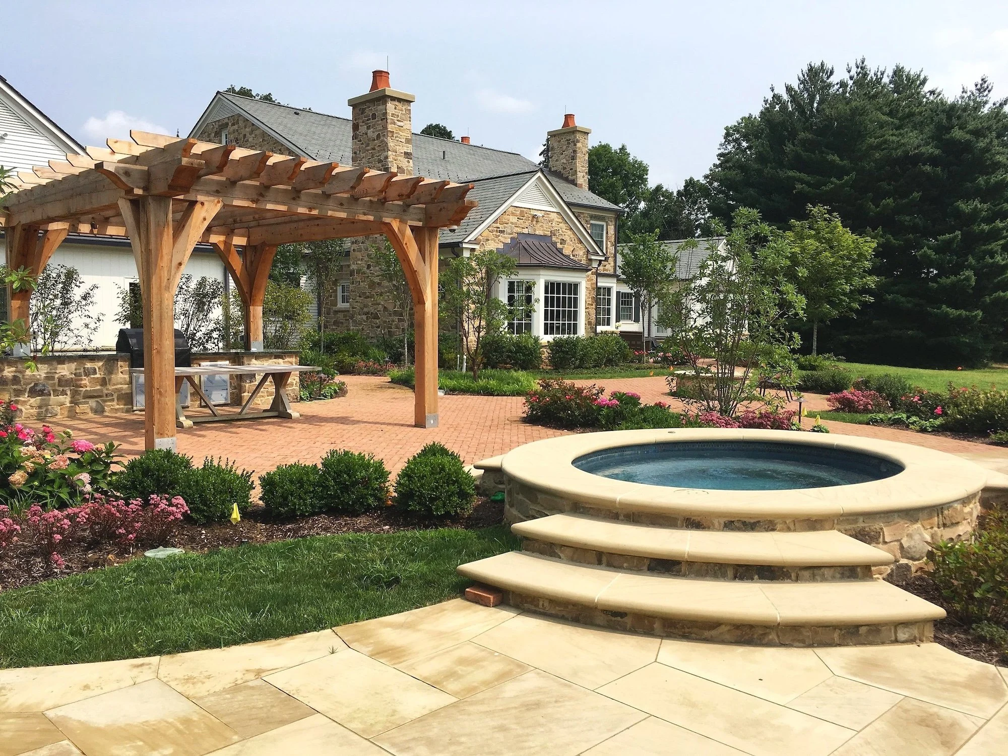 A backyard with a stone house, a wooden pergola, a hot tub, paved walkways, green grass, and landscaped flower beds.