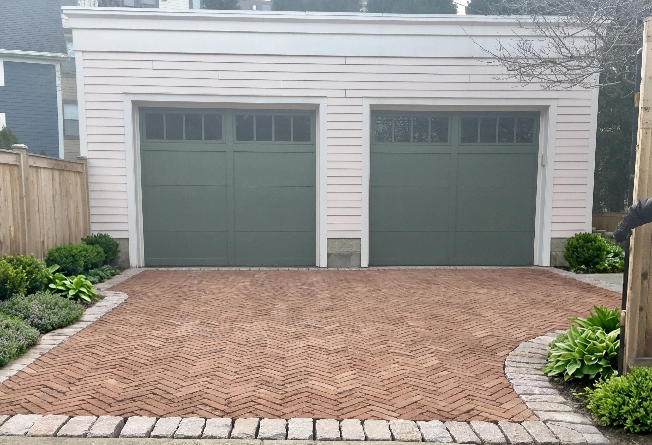 A paved driveway with brick pattern in front of a two-car garage attached to a house. The house has light pink siding, and there are plants and bushes along the sides of the driveway, with a wooden fence on the left and right sides.