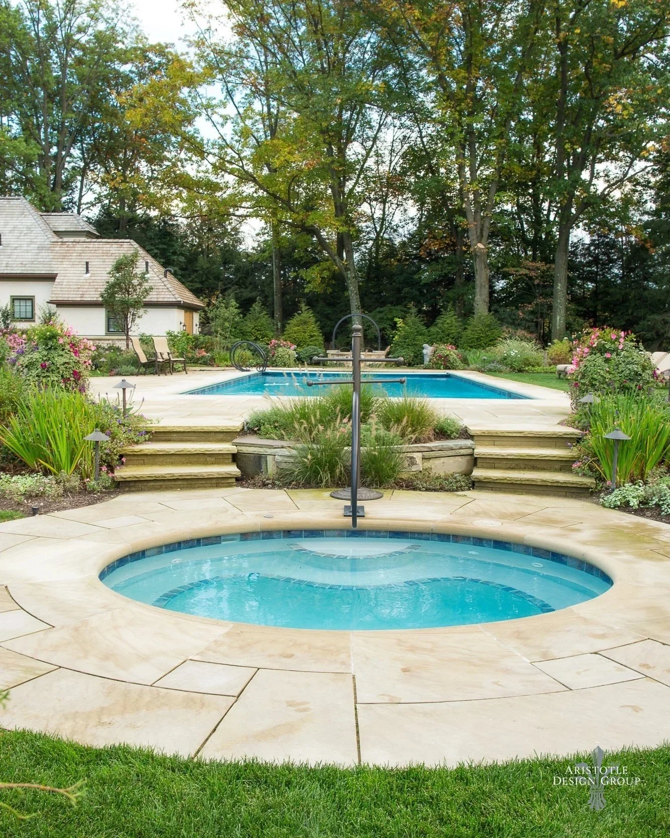A backyard with a swimming pool and hot tub surrounded by lush garden plants, trees, and a paved patio, with a house and tall trees in the background.