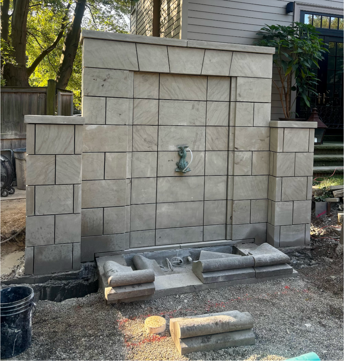 Partial view of a stone outdoor water fountain under construction with a small dragon or animal figure with a spout in the center, surrounded by beige stone blocks, on a gravel ground with some construction materials in front.