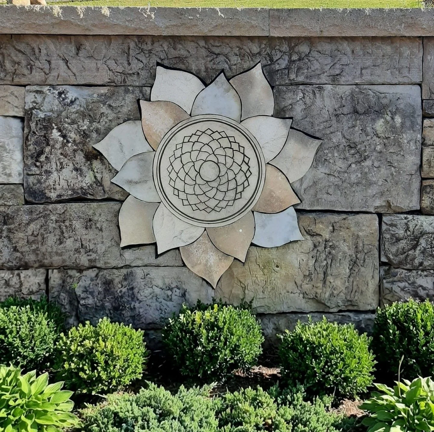 Decorative stone wall with a lotus flower design embedded in it, surrounded by green bushes at the bottom.