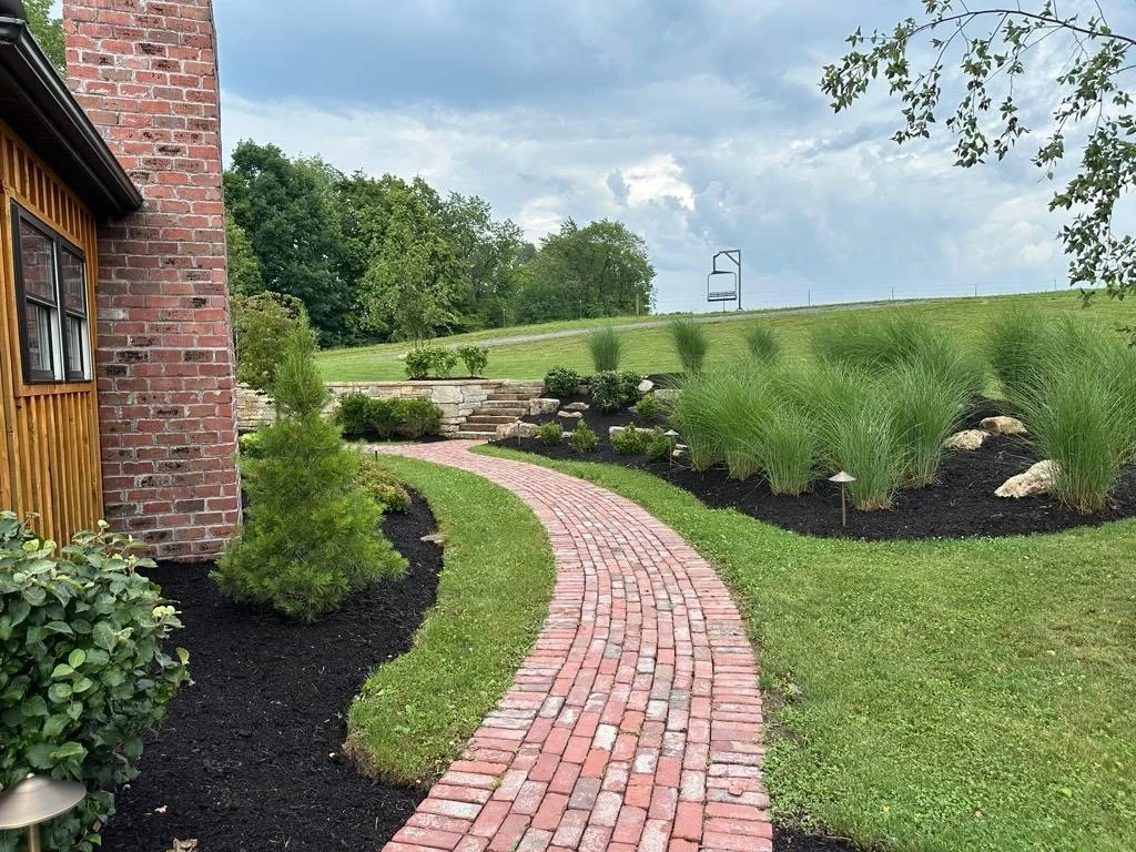 Brick pathway leading away from a house with brick and wood siding, surrounded by landscaped garden with bushes, grass, rocks, and ornamental grass, and a cloudy sky in the background.