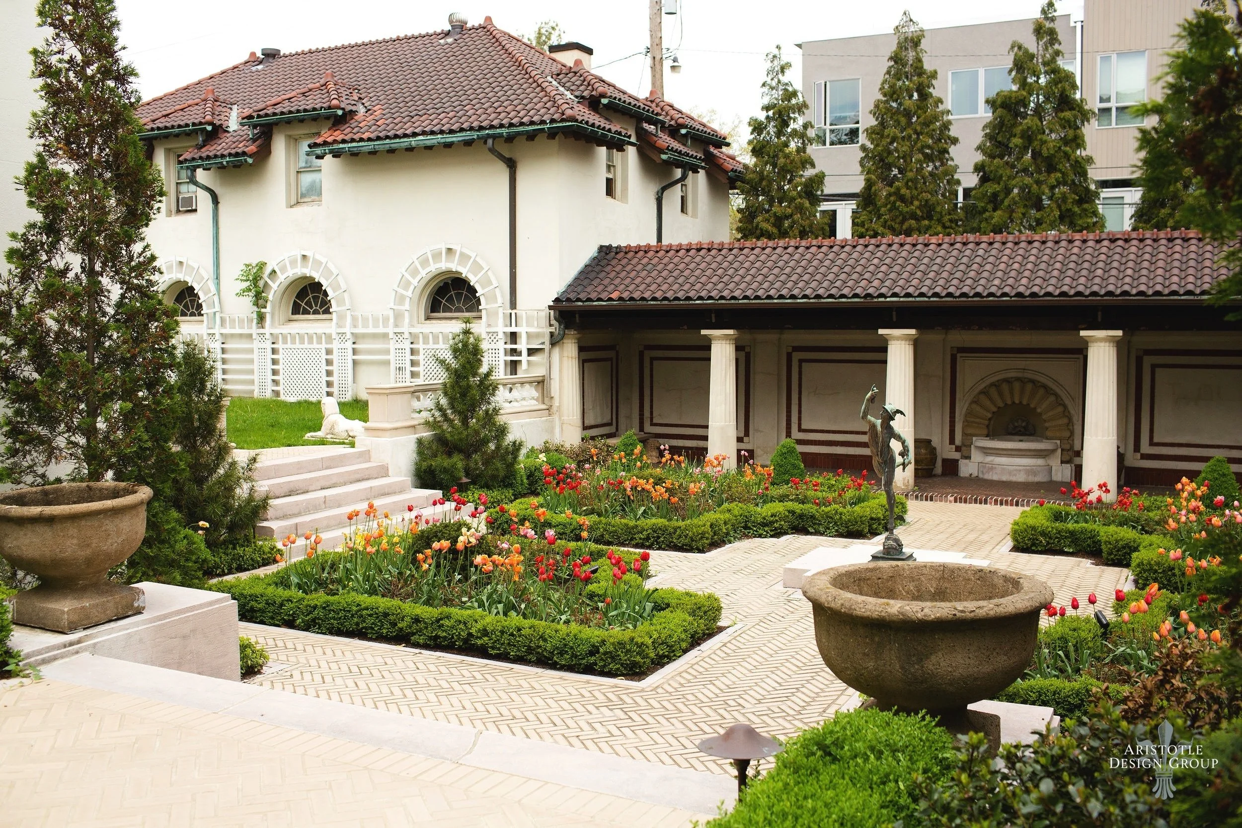 A landscaped garden with blooming tulips and trimmed bushes, stone planters, a statue of a woman, and a fountain, with elegant buildings and trees in the background.