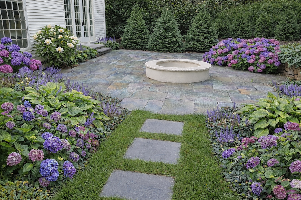 A landscaped backyard garden with purple and pink hydrangeas, green bushes, and a stone pathway leading to a circular stone fountain, adjacent to a house with white siding and large windows.