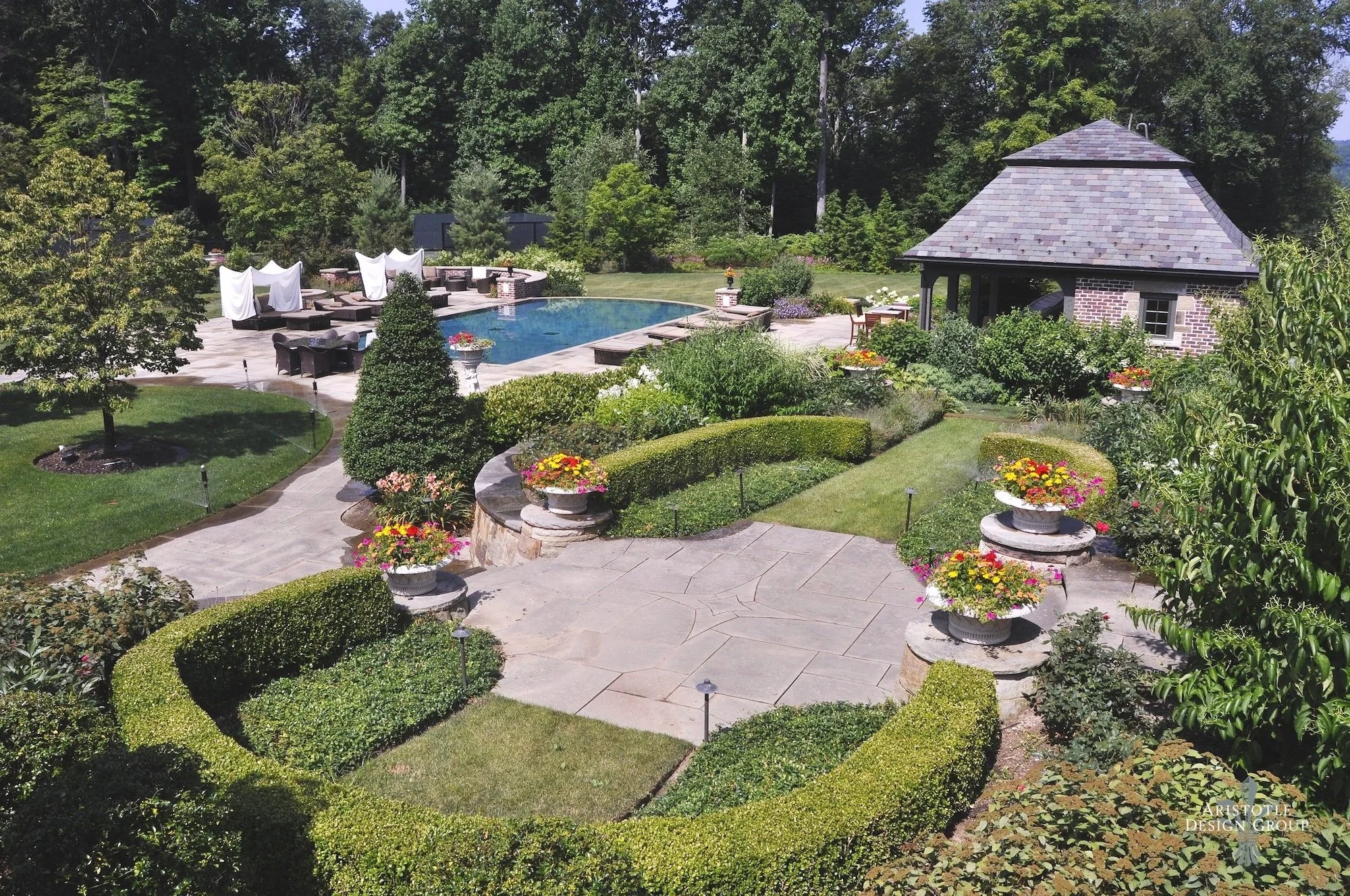 A luxurious backyard with a swimming pool, surrounded by lush green trees and well-manicured shrubs. There are several flower pots with colorful flowers, a stone patio, and a small brick pavilion with a shingled roof. Lounge chairs and umbrellas are 