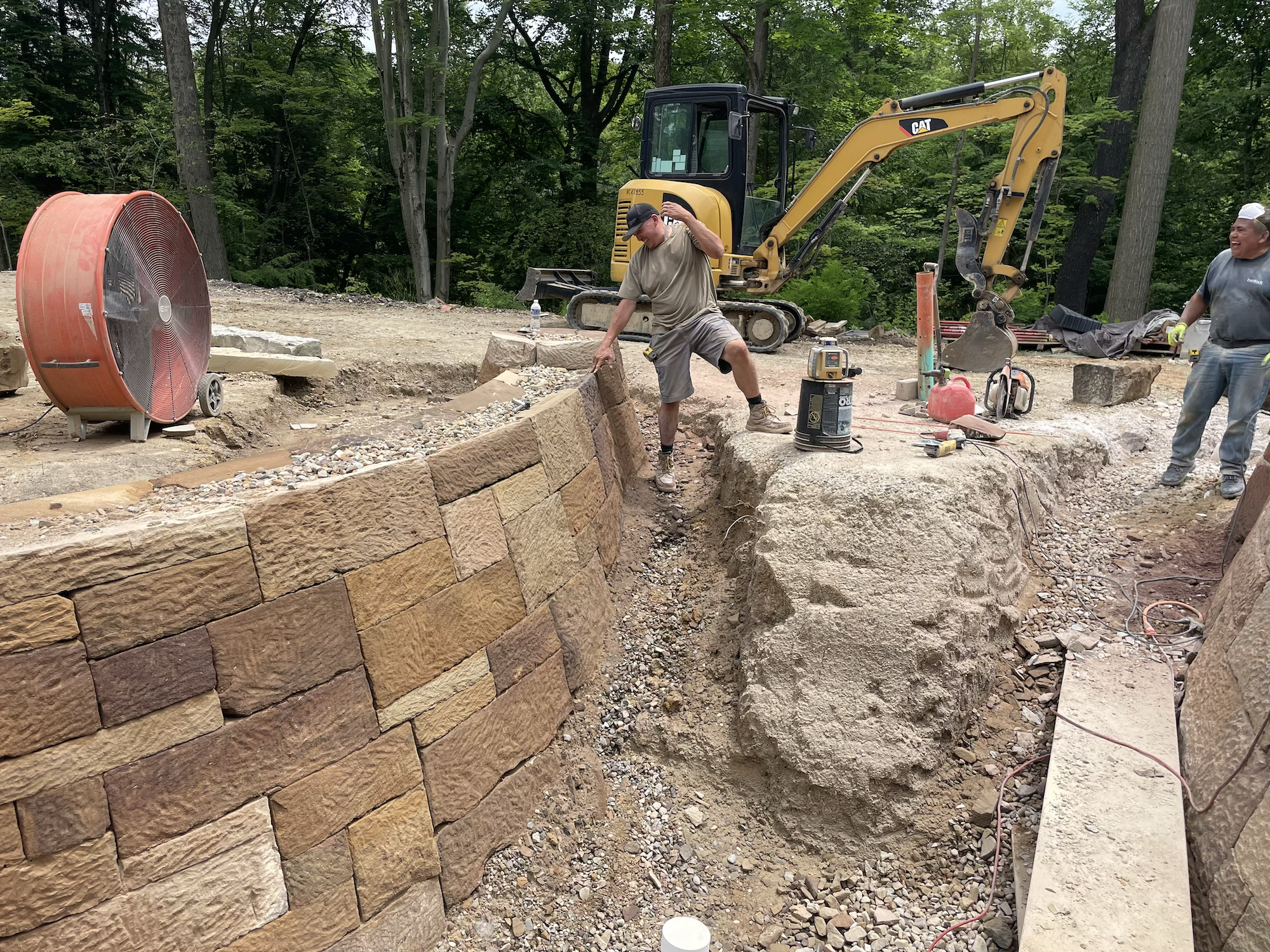 Two workers building a stone retaining wall in a forested area. One worker is placing a stone, while the other looks on, with construction equipment and a large fan nearby.