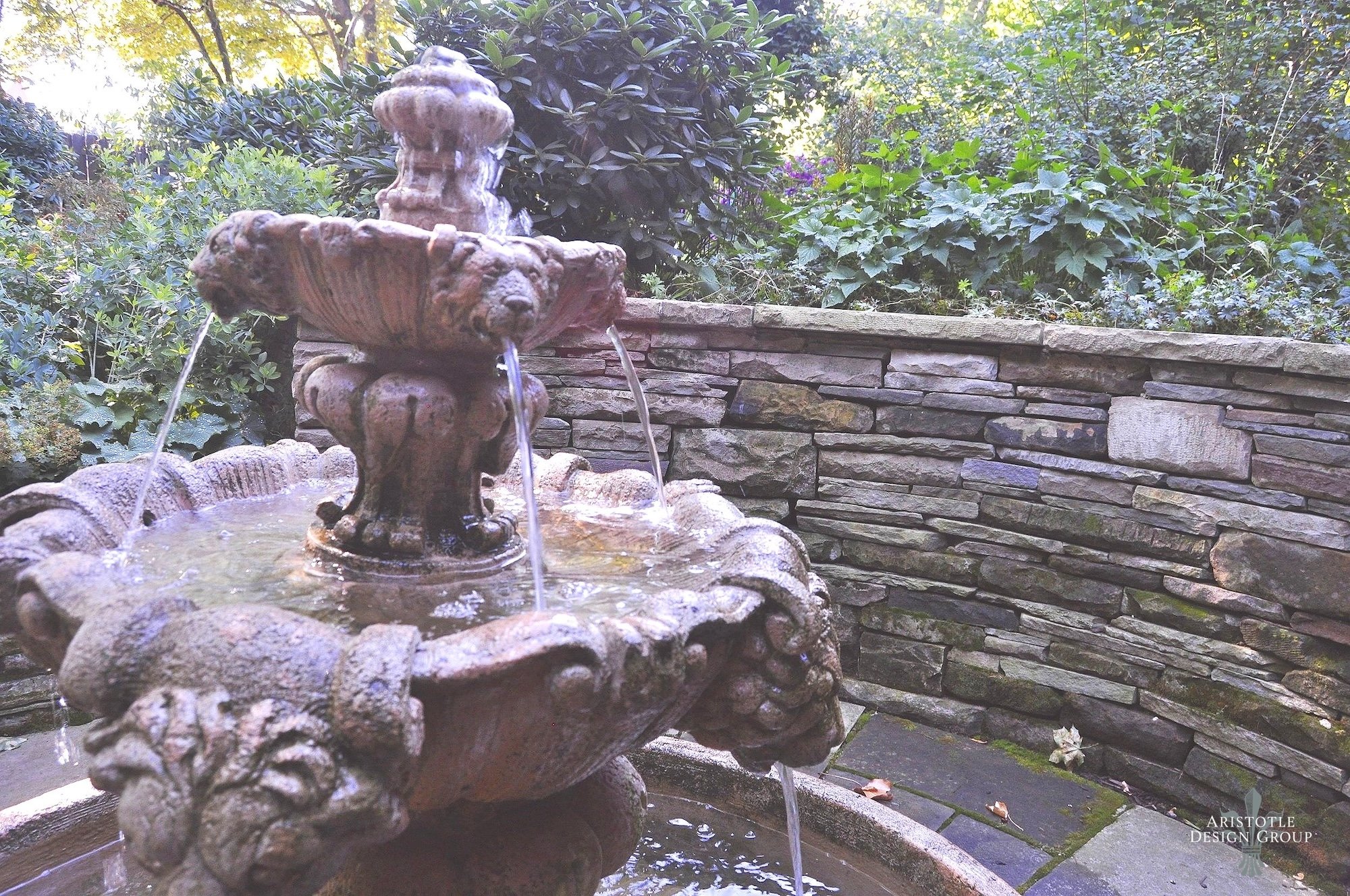 An ornate stone fountain with water cascading from multiple tiers in an outdoor garden setting, surrounded by lush green plants and a stone brick wall.