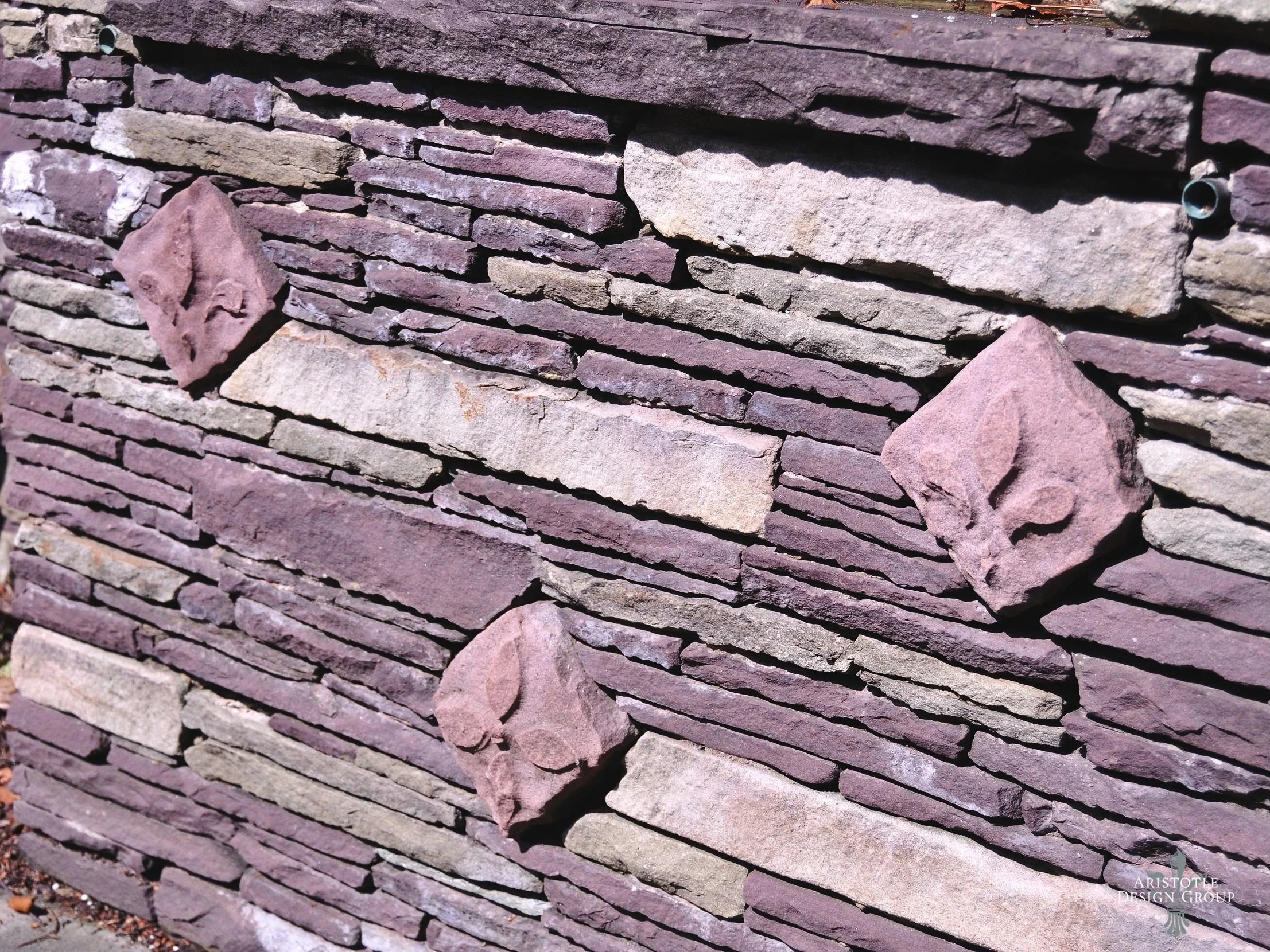 Close-up of a stone wall with horizontal layered flat stones. Three pinkish stones with carved skull designs are embedded in the wall, arranged vertically. The wall has a textured, natural appearance.