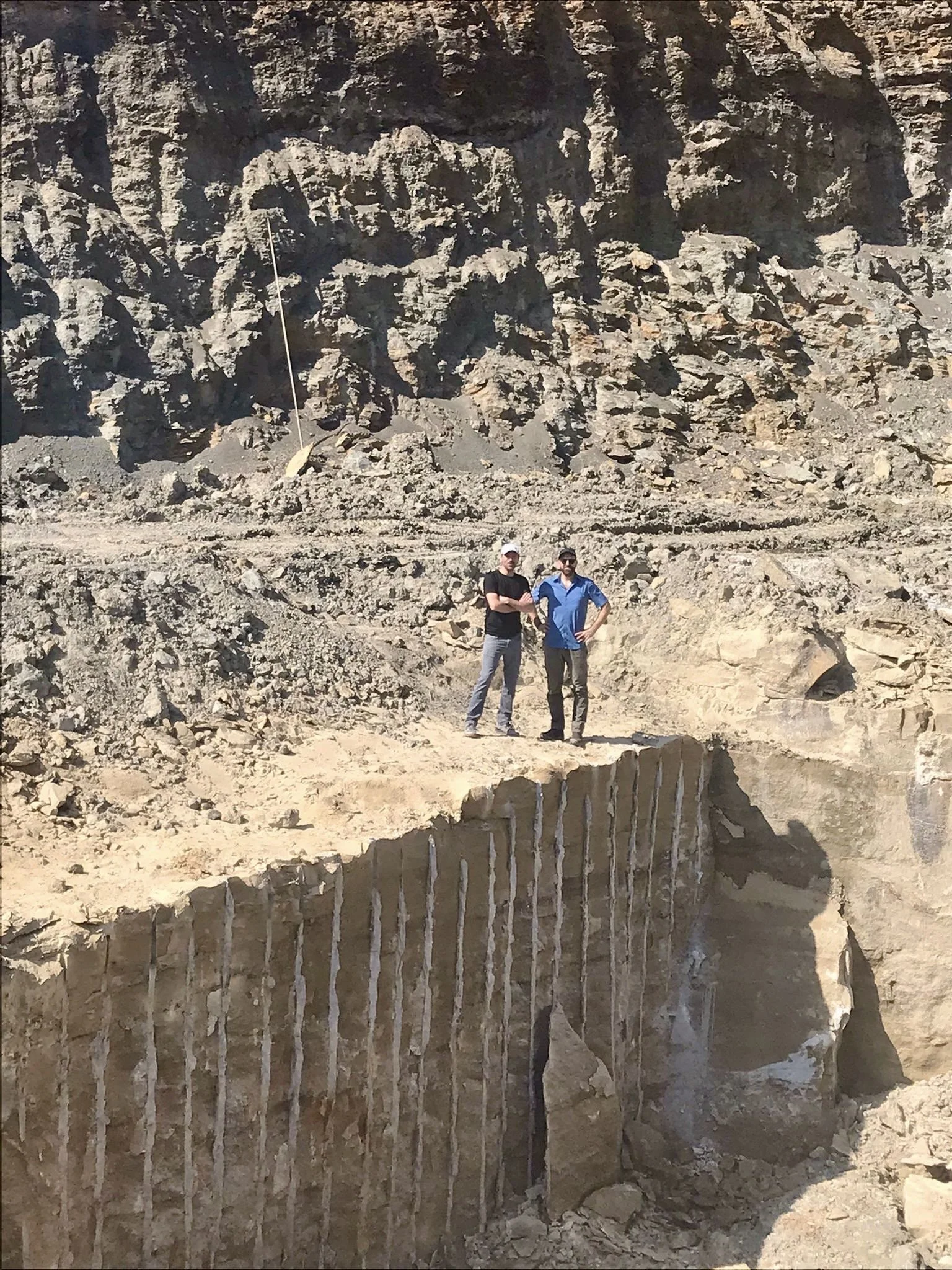 Two men stand together in a rocky desert landscape with a large cliff behind them. The cliff has visible layers of sediment and a crack with stalactites hanging down. One man is wearing a black shirt and gray pants, while the other is in a blue shirt