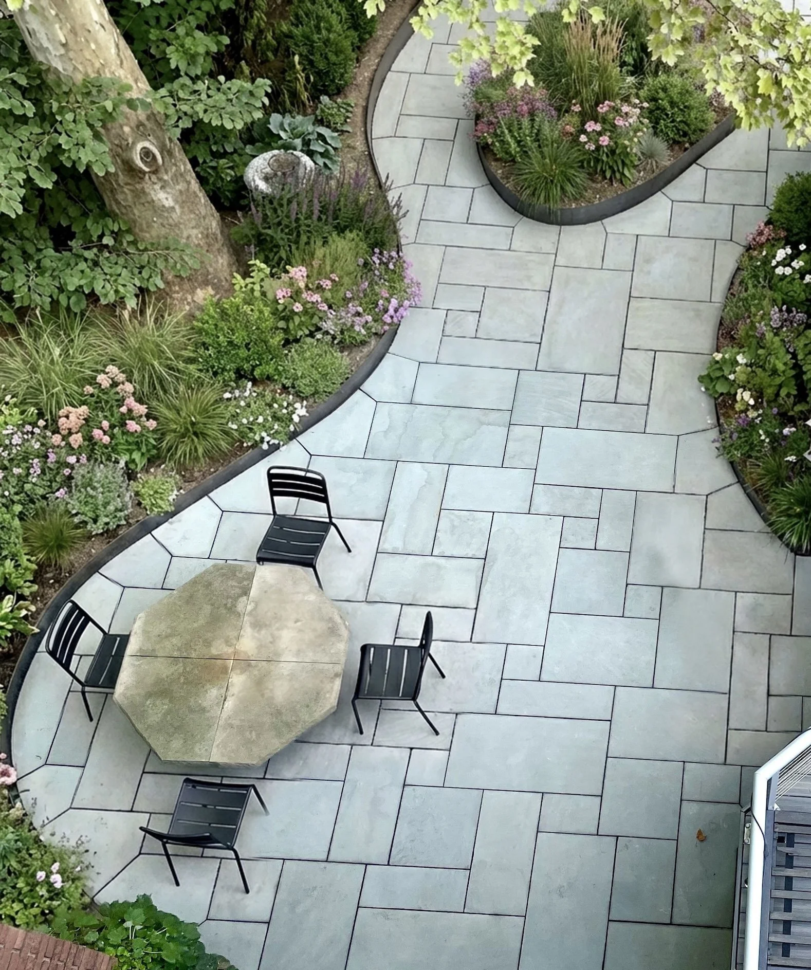 Overhead view of a patio with light-colored paving stones, garden beds with various plants and flowers, and a table with four black chairs.