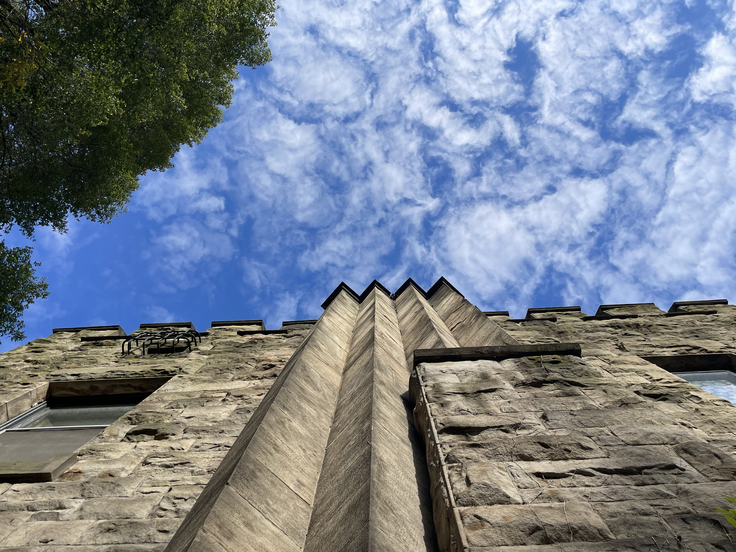 Upward view of a stone building with a corner trim at the top, with a background of blue sky and scattered clouds, and trees on the left side.