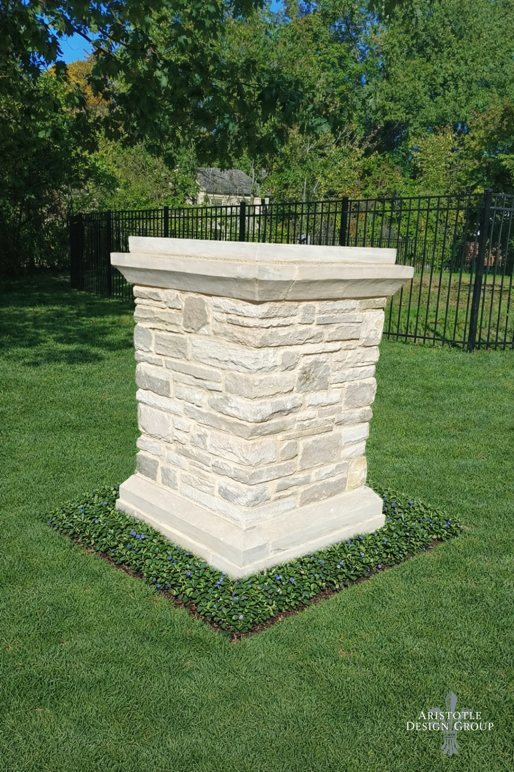 A stone pedestal with a square base and decorative crown molding on top, situated on a well-manicured lawn with a border of small green plants with purple flowers, surrounded by trees and a black metal fence.