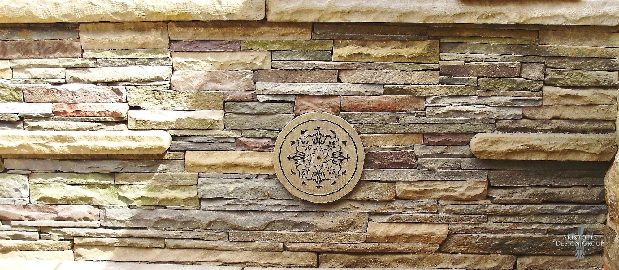 A stone wall with various sizes and colors of stacked rocks and a decorative circular emblem with intricate black patterns in the center. The logo of Aristotle Design Group is in the lower right corner.